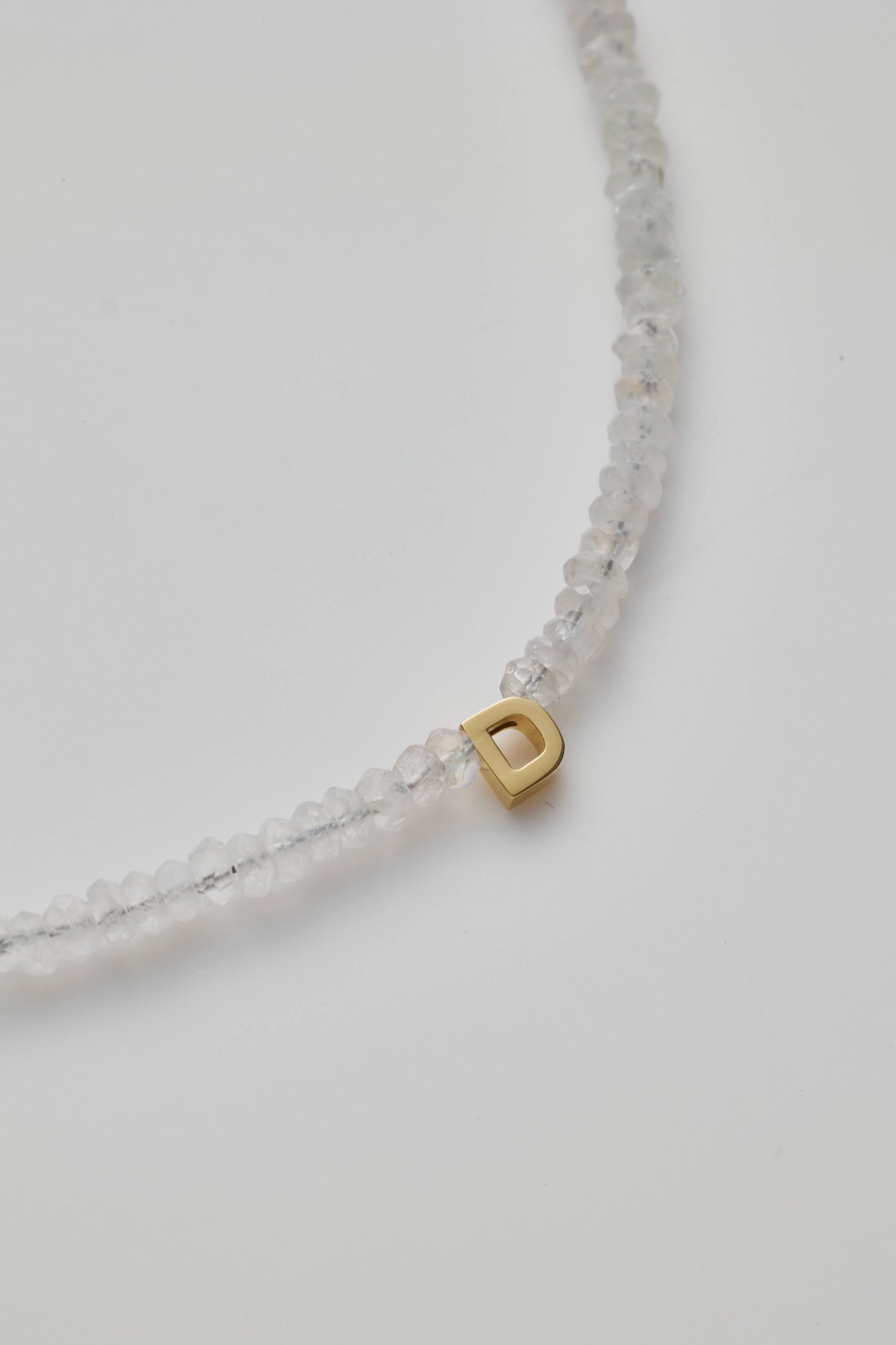 The One Letter Bracelet in Rainbow Moonstone and 14k Gold features small, translucent beads with a single 18k solid gold bead displaying the letter "D." This elegant bracelet includes a gold clasp closure and can be customised to fit different sizes. The product is showcased against a plain white background.