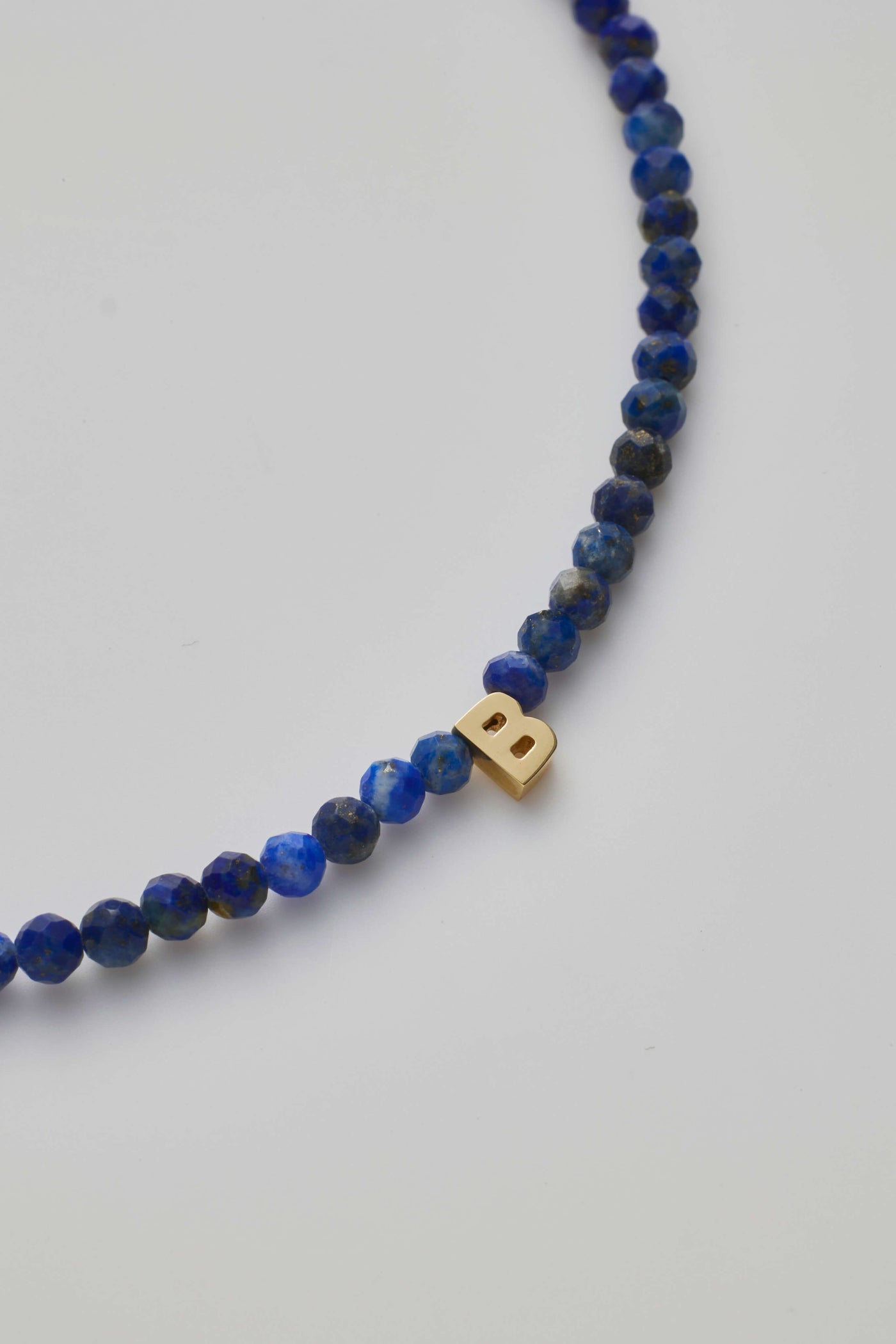 A close-up of the One Letter Bracelet in Lapis Lazuli and 14k Gold, showcasing small, round, faceted blue beads. At its centre is a single 14k solid gold bead shaped like the letter "B." The bracelet is photographed against a plain white background.