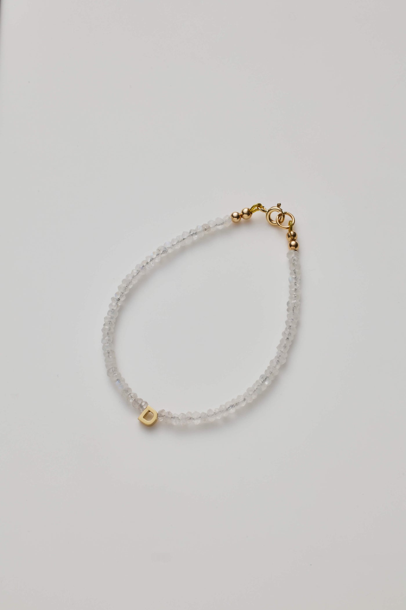 The One Letter Bracelet in Rainbow Moonstone and 14k Gold features small, translucent beads with a single 18k solid gold bead displaying the letter "D." This elegant bracelet includes a gold clasp closure and can be customised to fit different sizes. The product is showcased against a plain white background.