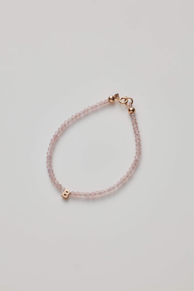 The One Letter Bracelet in Rose Quartz and 14k Rose Gold features small, round rose quartz beads and a gold lobster clasp. A single 14k gold initial charm, the letter "B," is threaded onto the beads. The bracelet is available in custom sizes, arranged in a simple, curved shape on a white background.