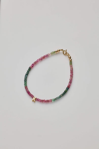 The One Letter Bracelet in Tourmaline and 14k Gold showcases delicate, small multicoloured beads arranged in a beautiful gradient from pink to green. The piece is elegantly finished with an 14k solid gold clasp and a charming letter "K" charm. Displayed on a white background in an oval shape, the bracelet is available in custom sizes.