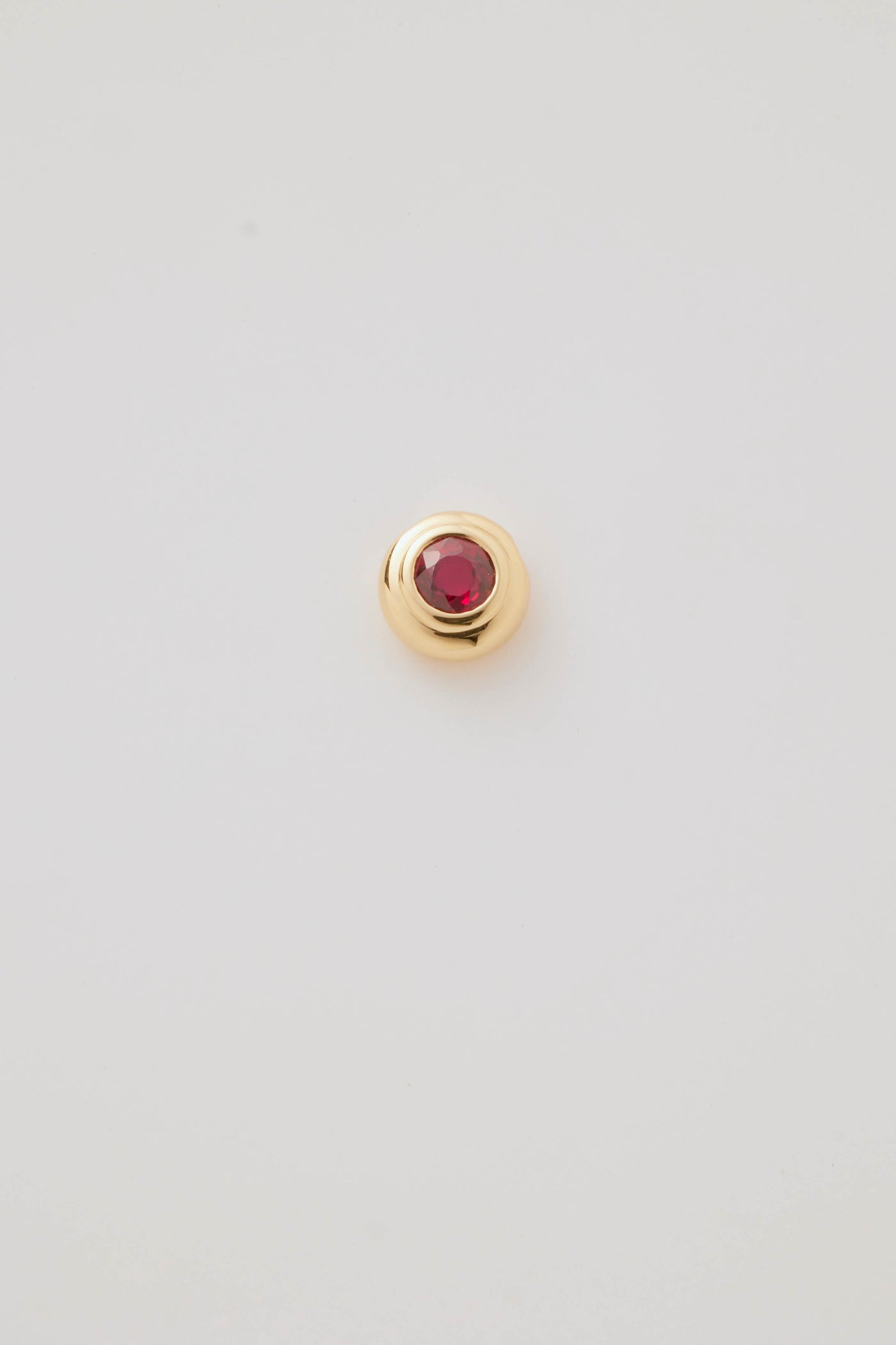 Close-up image of a July Birthstone Charm featuring a vibrant ruby gemstone in a polished gold setting. Photographed on a pale grey background. 