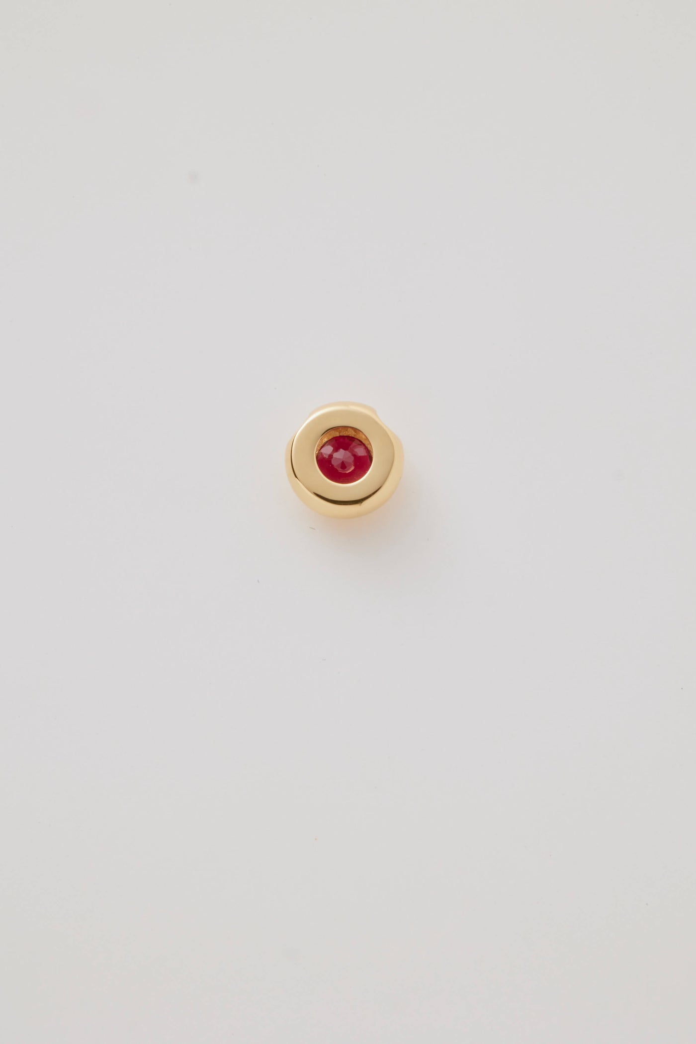 The reverse side view of a July Birthstone Charm featuring a vibrant ruby gemstone in a polished gold setting. Photographed on a pale grey background. 