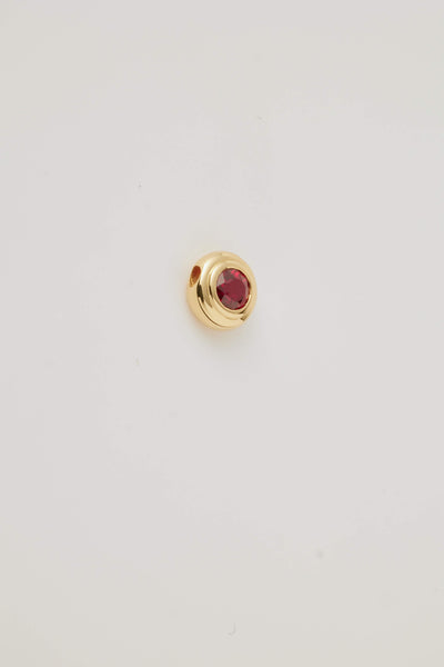 Side view image of a July Birthstone Charm featuring a vibrant ruby gemstone in a polished gold setting. Photographed on a pale grey background. 