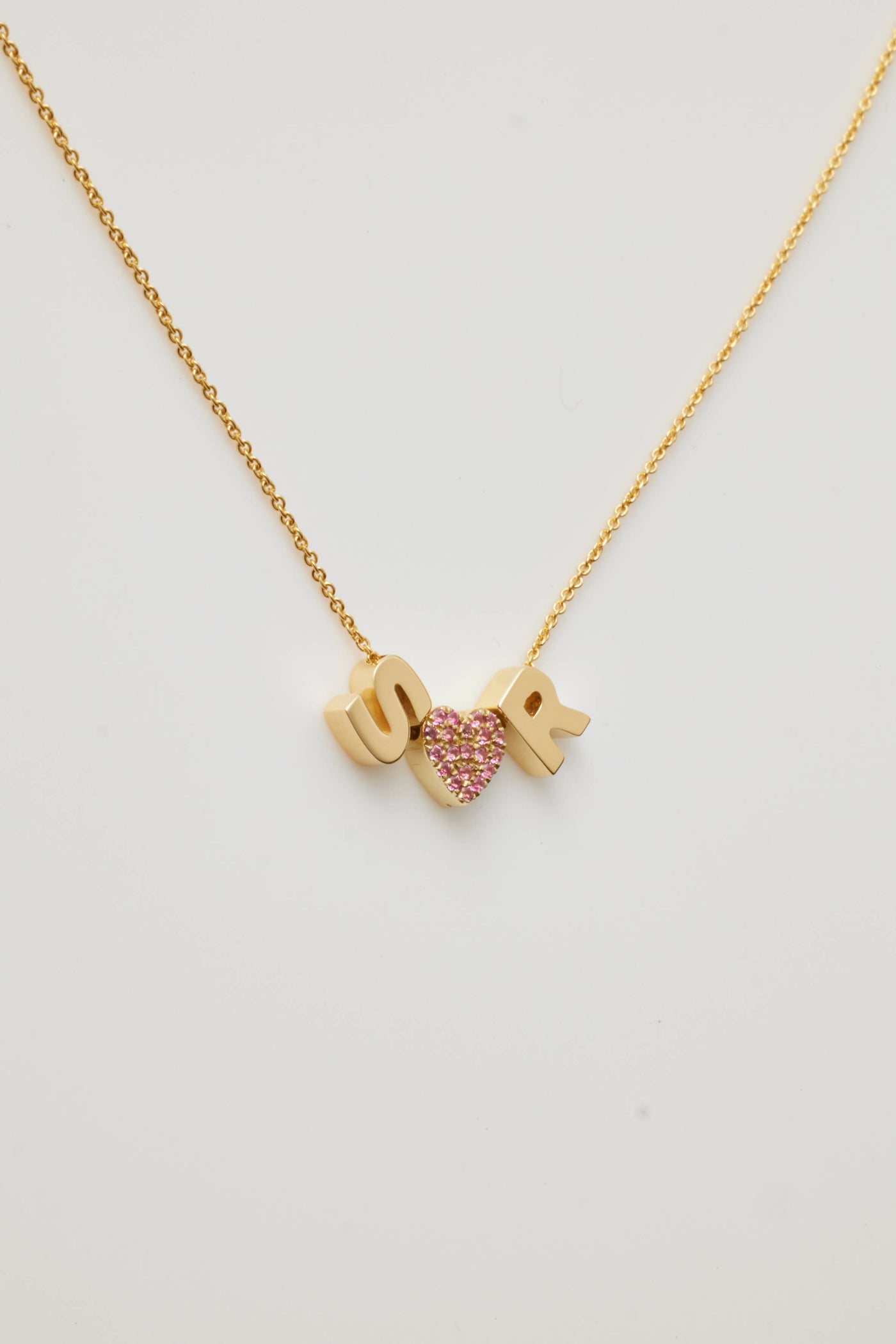 A delicate Two Letters and Pink Sapphire Heart Necklace in 18k Gold features three hanging pendants with two gold letters "SR." and "heart "the heart is set with vibrant hued pink sapphires, encrusted in gemstones. The necklace lies gracefully on a white background.