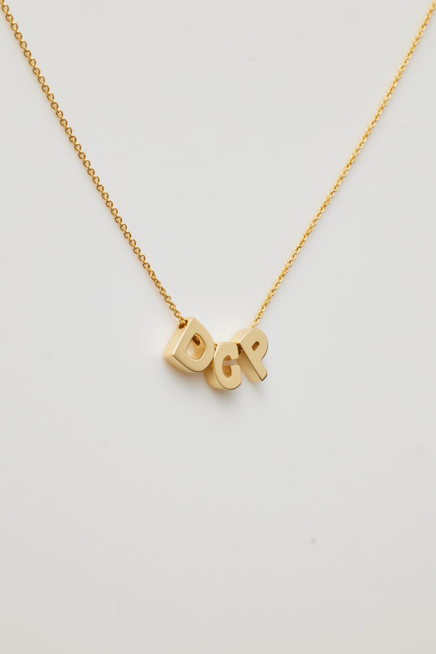 The Three Letters Necklace in 18k Gold is a delicate piece featuring three polished gold letters as the pendants. This sophisticated necklace, with its fine chain, gracefully displays the initials "DGP," which hang prominently at the centre, casting a slight shadow on any light background.