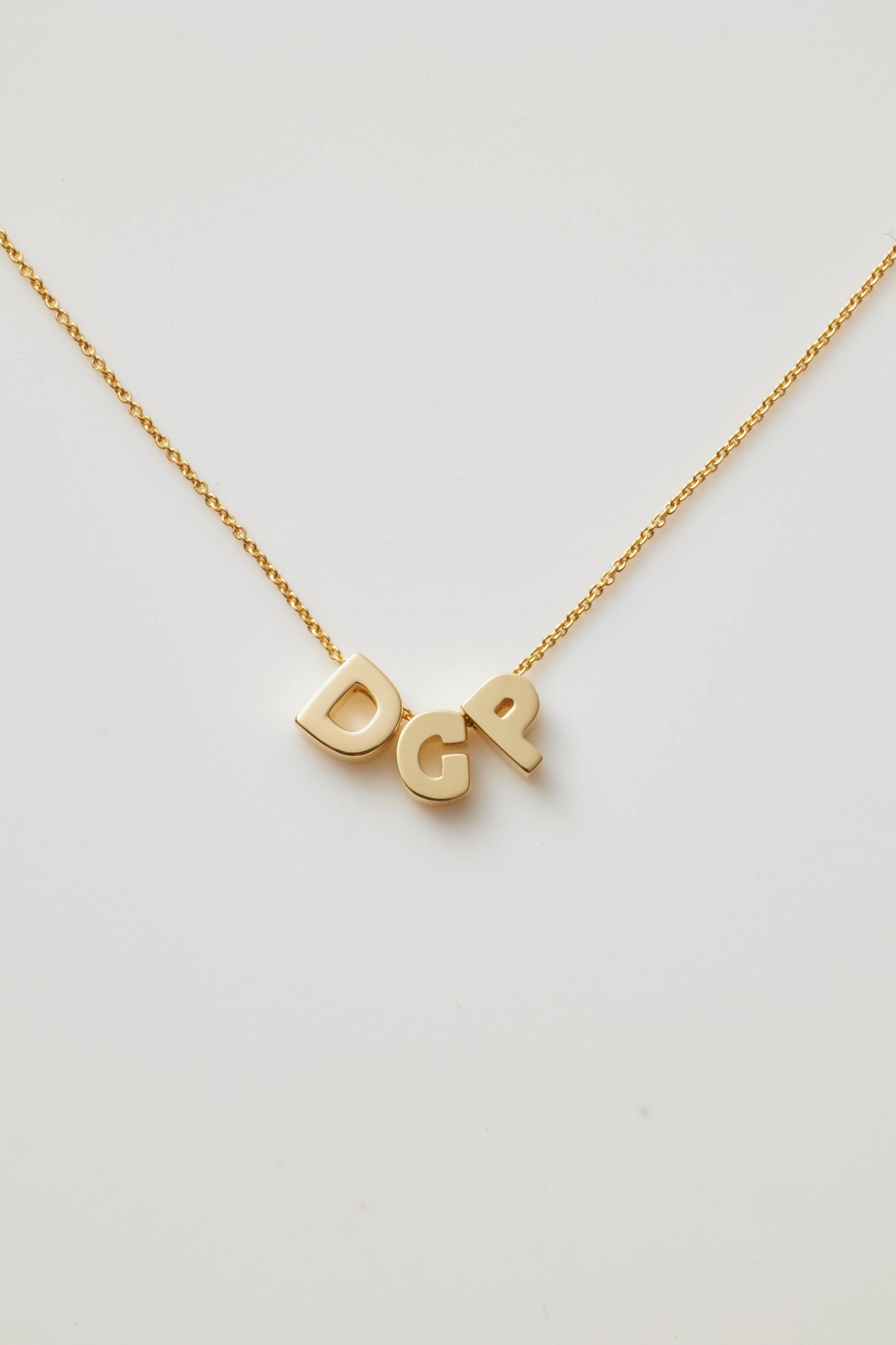 The Three Letters Necklace in 18k Gold is a delicate piece featuring three polished gold letters as the pendants. This sophisticated necklace, with its fine chain, gracefully displays the initials "DGP," which hang prominently at the centre, casting a slight shadow on any light background.