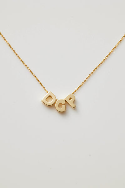 The Three Letters Necklace in 18k Gold is a delicate piece featuring three polished gold letters as the pendants. This sophisticated necklace, with its fine chain, gracefully displays the initials "DGP," which hang prominently at the centre, casting a slight shadow on any light background.