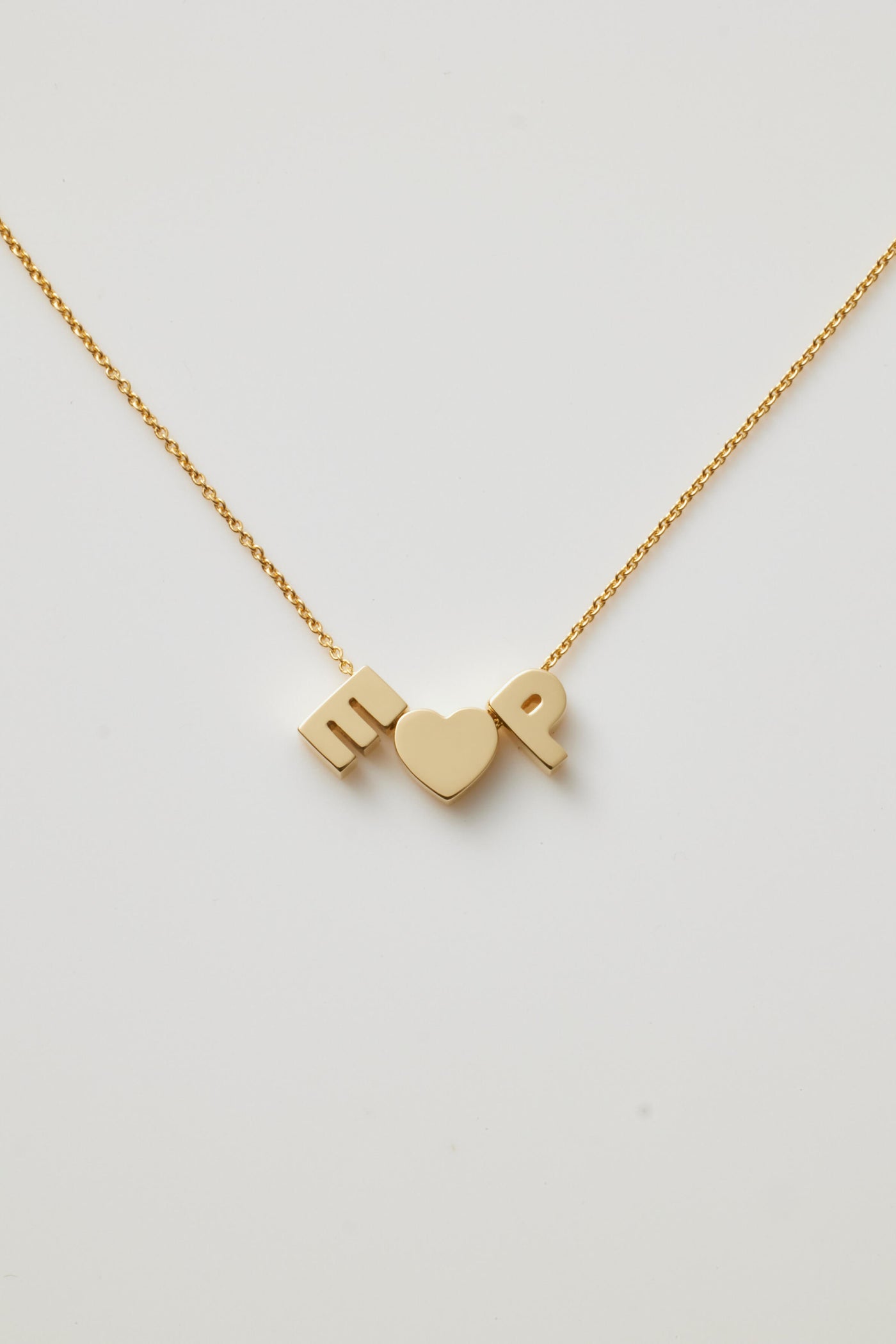 The Two Letters and Heart Necklace in 18k Gold boasts a delicate chain adorned with three exquisite charms: a capital letter "E" a heart symbol centrally placed, and a capital letter "P." Made from 18k solid gold, this beautiful piece is displayed against a simple, light-coloured backdrop and features an openable connector clasp for effortless wear.