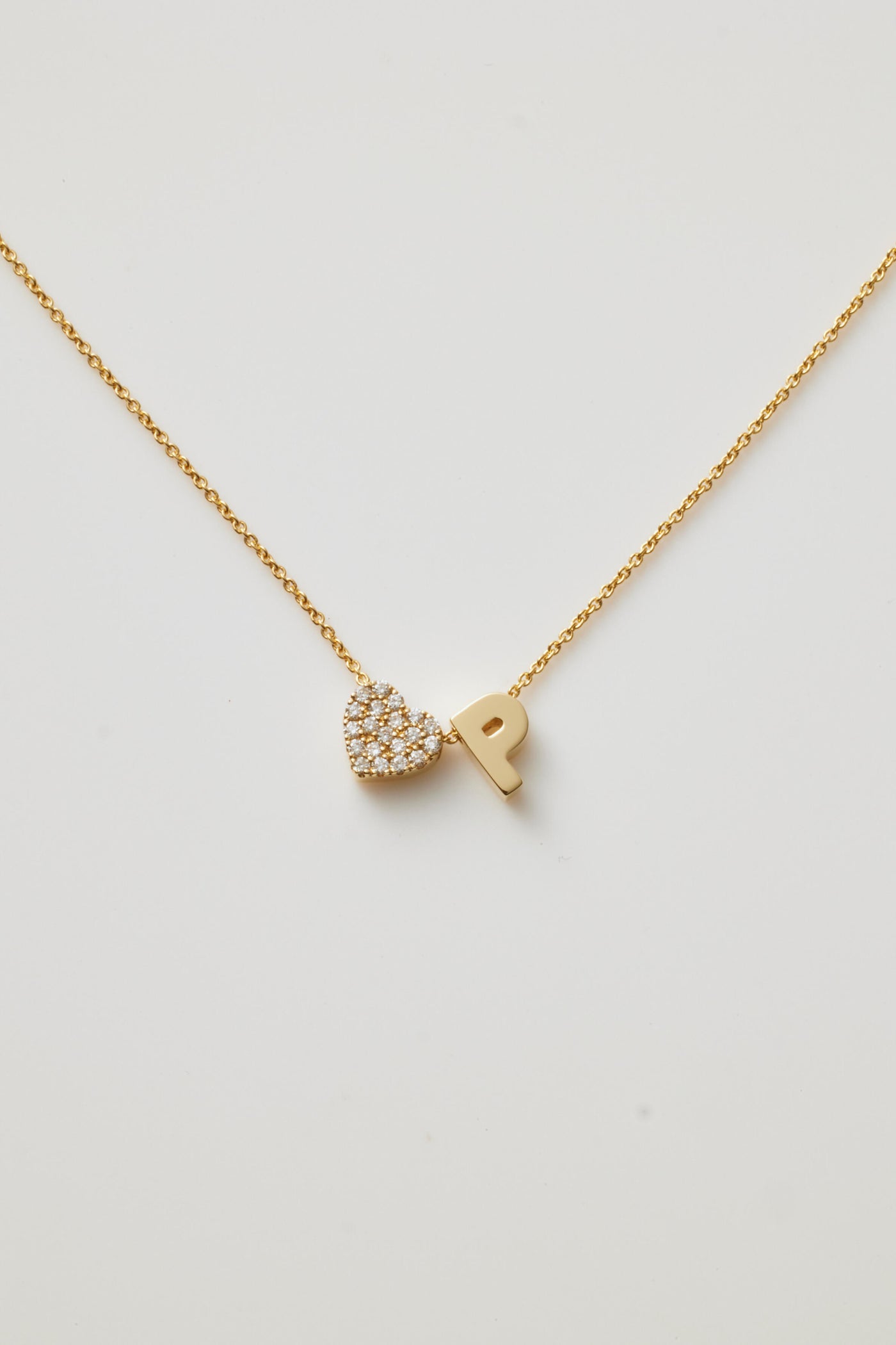 A gold necklace featuring a delicate chain with two charms: a sparkling, diamond-studded heart and a bold gold letter "P," positioned side by side.