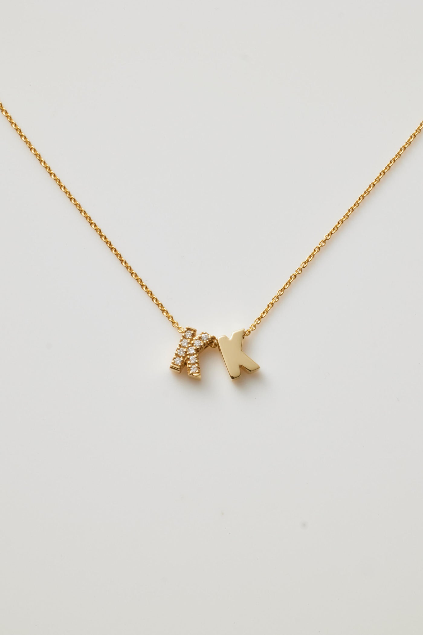 The Two Letters Necklace in Diamonds and 18k Gold showcases a minimalist and elegant design with two hanging pendants: a solid 18k gold "K" and a gold "K" encrusted with sparkling diamonds. This stunning necklace shines beautifully against a grey background.