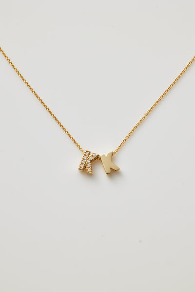 The Two Letters Necklace in Diamonds and 18k Gold showcases a minimalist and elegant design with two hanging pendants: a solid 18k gold "K" and a gold "K" encrusted with sparkling diamonds. This stunning necklace shines beautifully against a grey background.