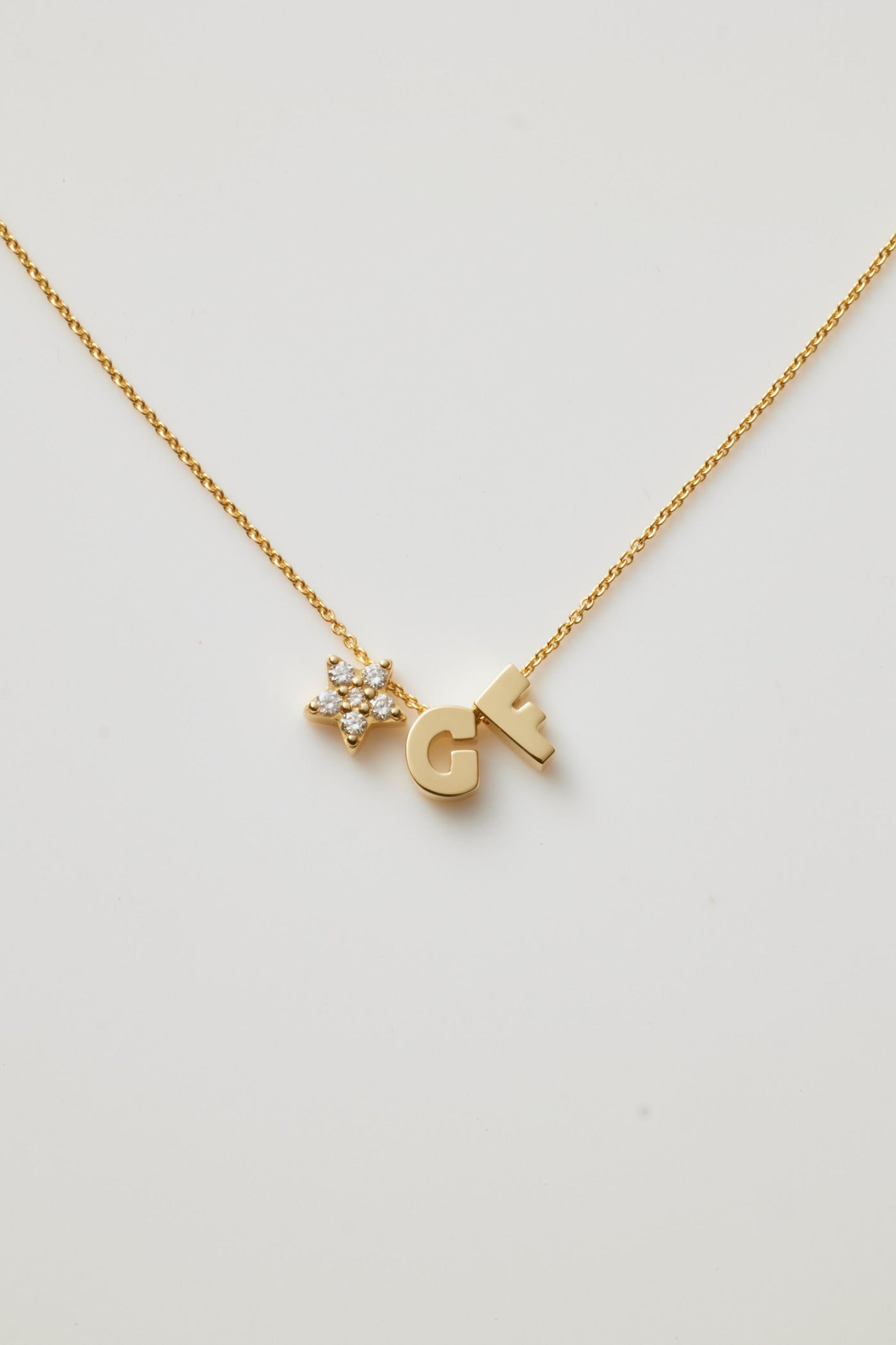 The "Two Letters and Diamond Star Necklace in 18k Gold" is a delicate piece showcasing two letter charms, "G" and "F," flanking a radiant star-shaped charm adorned with round brilliant diamonds. All are elegantly suspended from a fine gold chain against a white background.