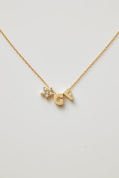 The "Two Letters and Diamond Star Necklace in 18k Gold" is a delicate piece showcasing two letter charms, "G" and "F," flanking a radiant star-shaped charm adorned with round brilliant diamonds. All are elegantly suspended from a fine gold chain against a white background.