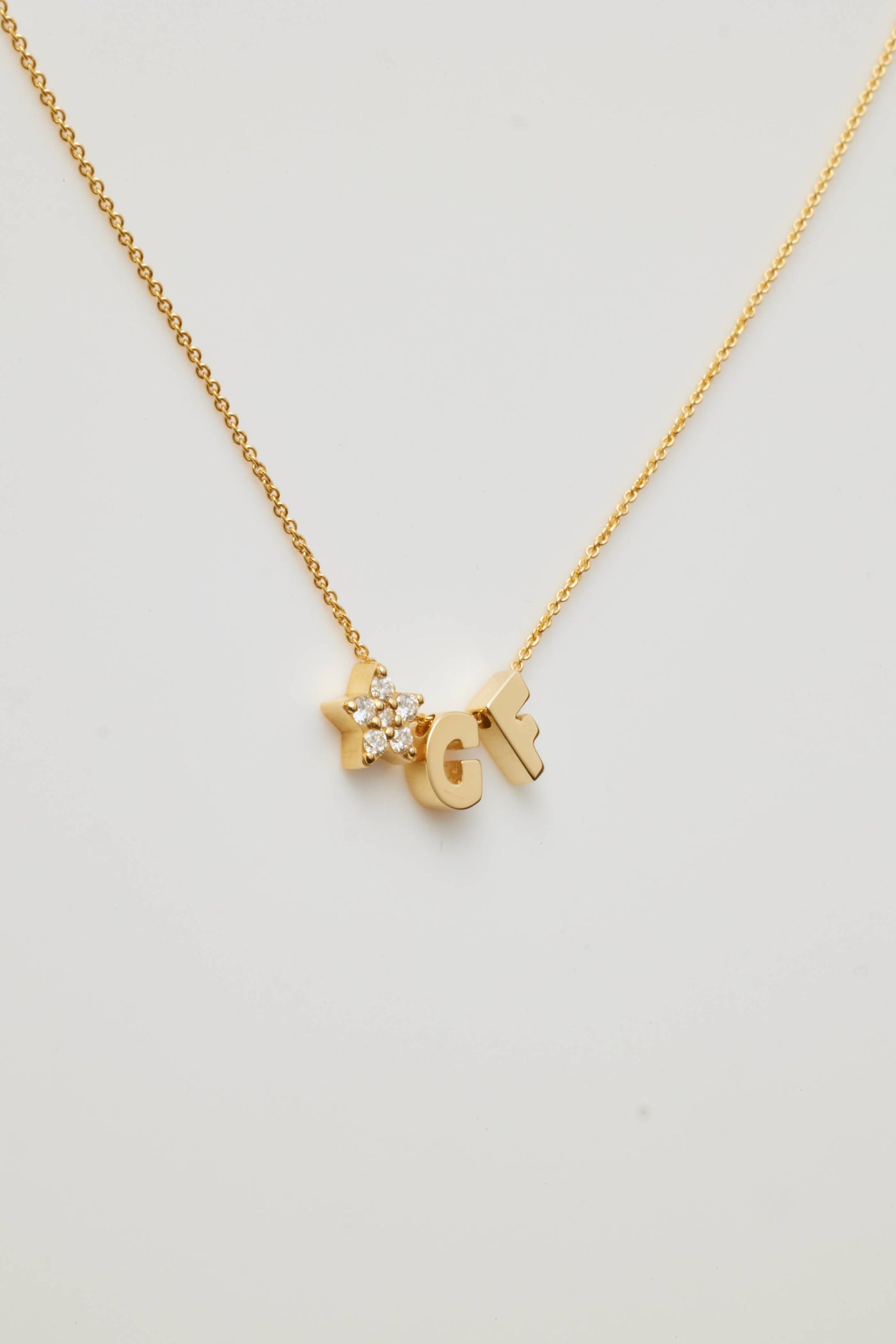 The "Two Letters and Diamond Star Necklace in 18k Gold" is a delicate piece showcasing two letter charms, "G" and "F," flanking a radiant star-shaped charm adorned with round brilliant diamonds. All are elegantly suspended from a fine gold chain against a white background.