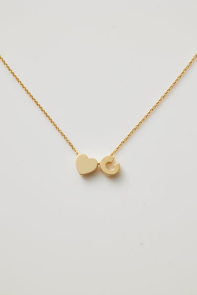 The One Letter and Heart Necklace in 18k Gold features a delicate solid gold chain adorned with two pendants: a heart and a "C". The heart and letter contribute to a simple, elegant design against a white background, exuding timeless charm.