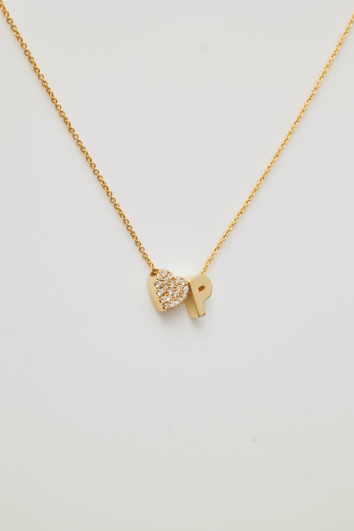 A gold necklace featuring a delicate chain with two charms: a sparkling, diamond-pave heart and a bold gold letter "P," positioned side by side.