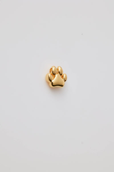 Side view of a polished gold dog paw charm with intricate detailing or raised paw pads and hole at the top for a chain to thread through. Background is a light grey colour.