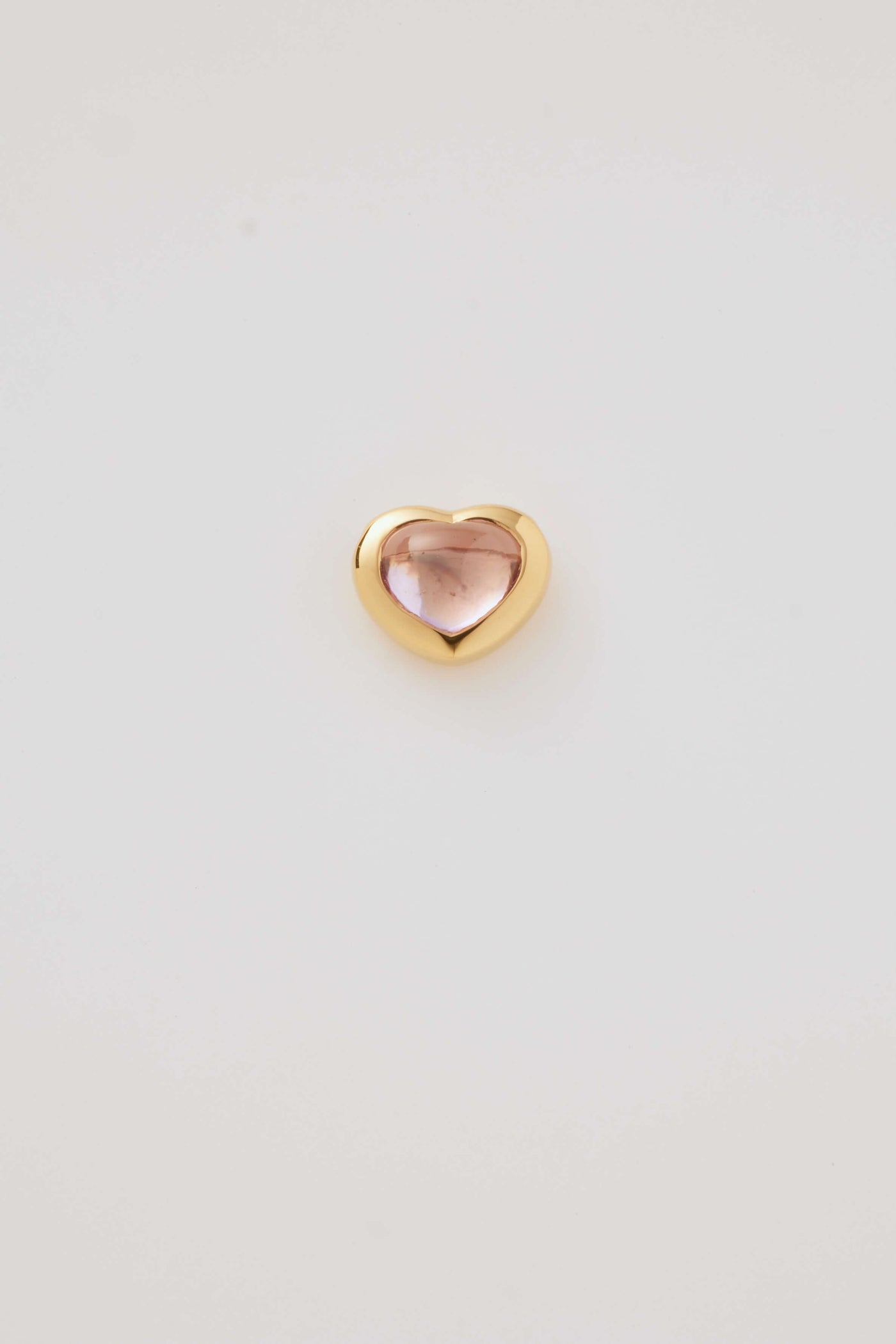 Front profile of a pink cabochon stone set in a polished gold heart frame, displayed on a light grey background. Elegant and timeless.