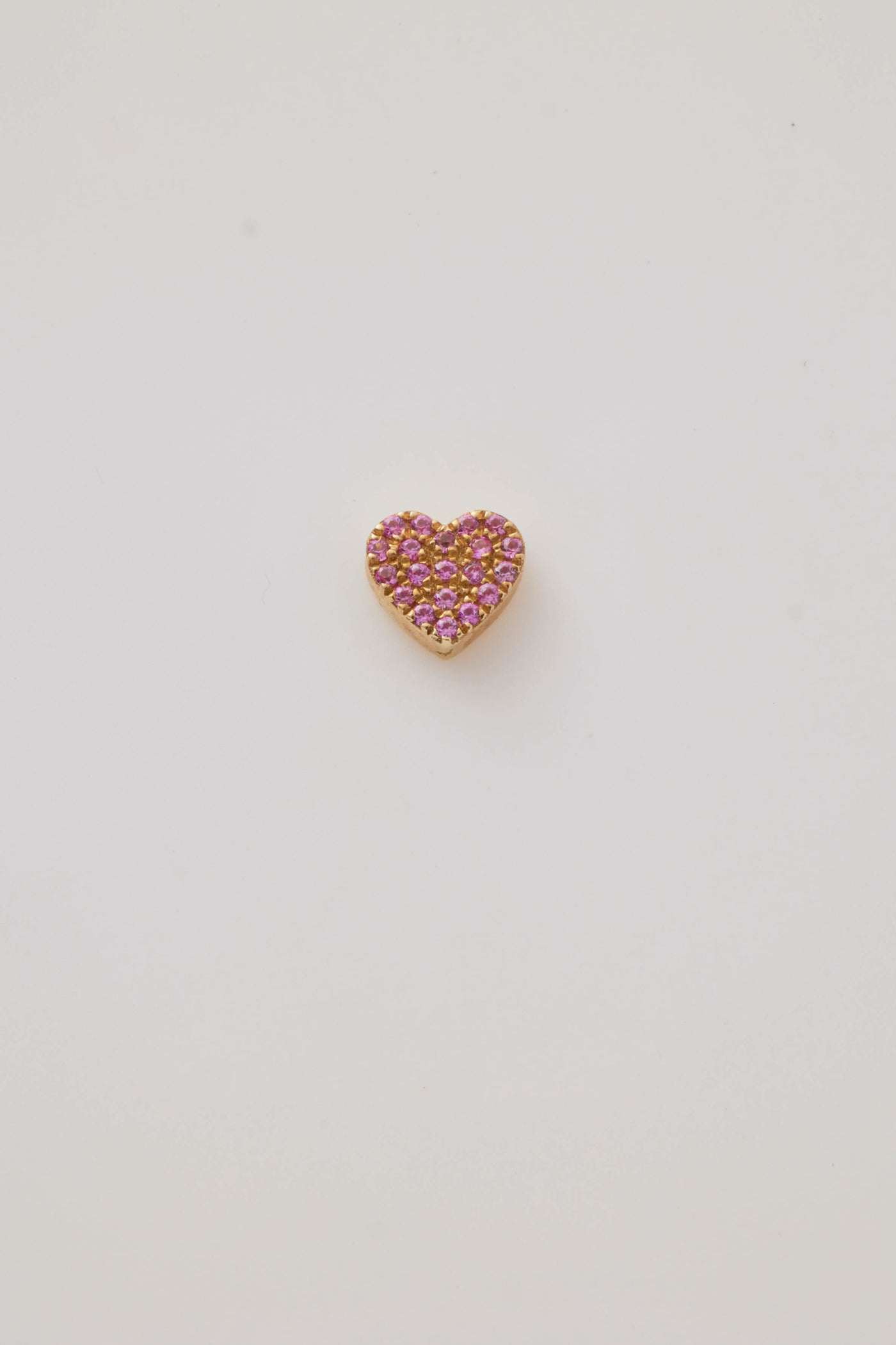 A solid gold miniature heart charm, elegantly adorned with vibrant pink sapphire gemstones. The tiny heart-shaped design is pavé-set with sparkling pink sapphires, creating a vivid, eye-catching effect. A small hole at the top allows the charm to be easily threaded through to a gold chain adding a touch of luxury and romance.