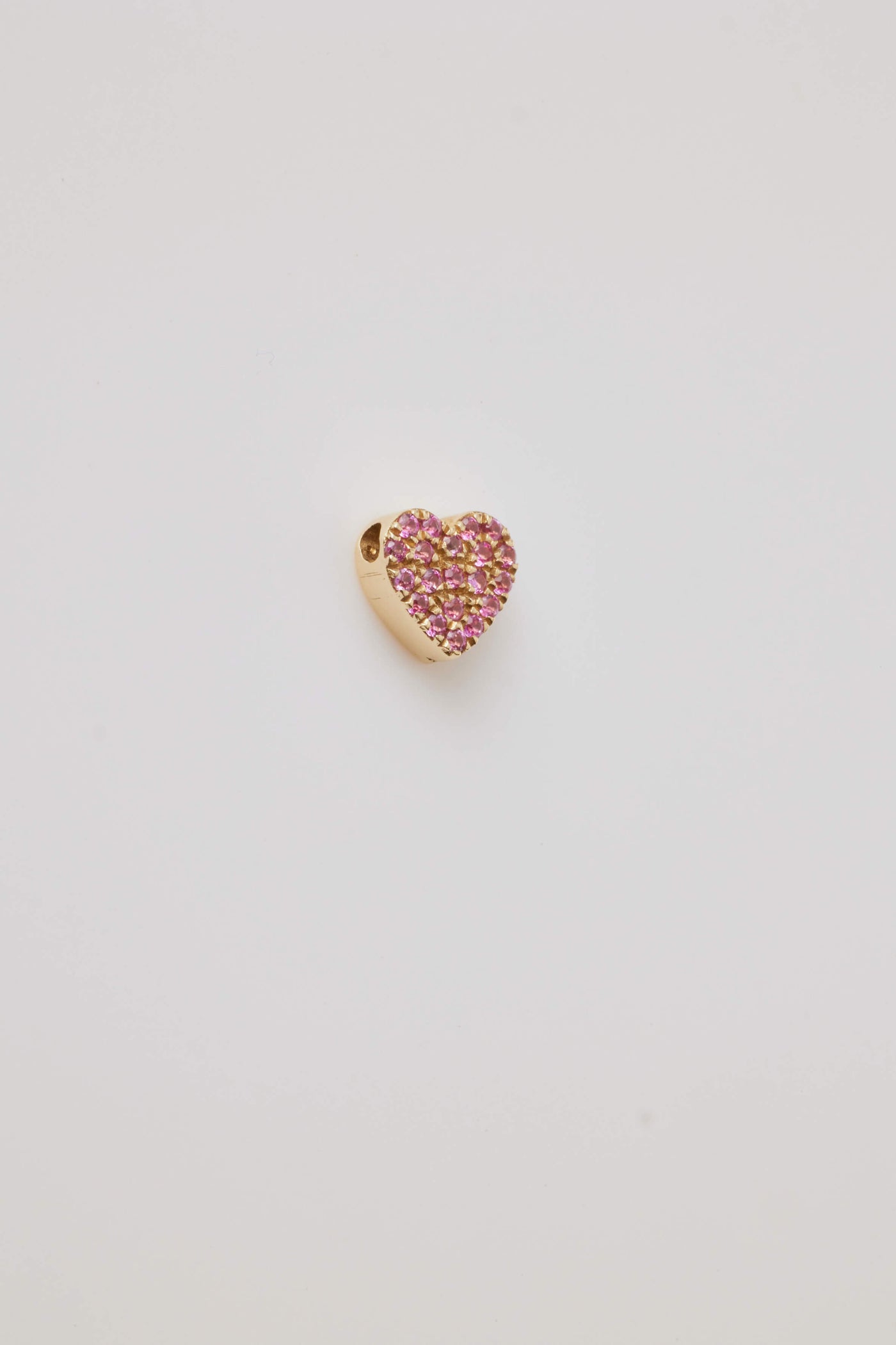 A side view of a solid gold miniature heart charm, elegantly adorned with vibrant pink sapphire gemstones. The tiny heart-shaped design is pavé-set with sparkling pink sapphires, creating a vivid, eye-catching effect. A small hole at the top allows the charm to be easily threaded through to a gold chain adding a touch of luxury and romance.