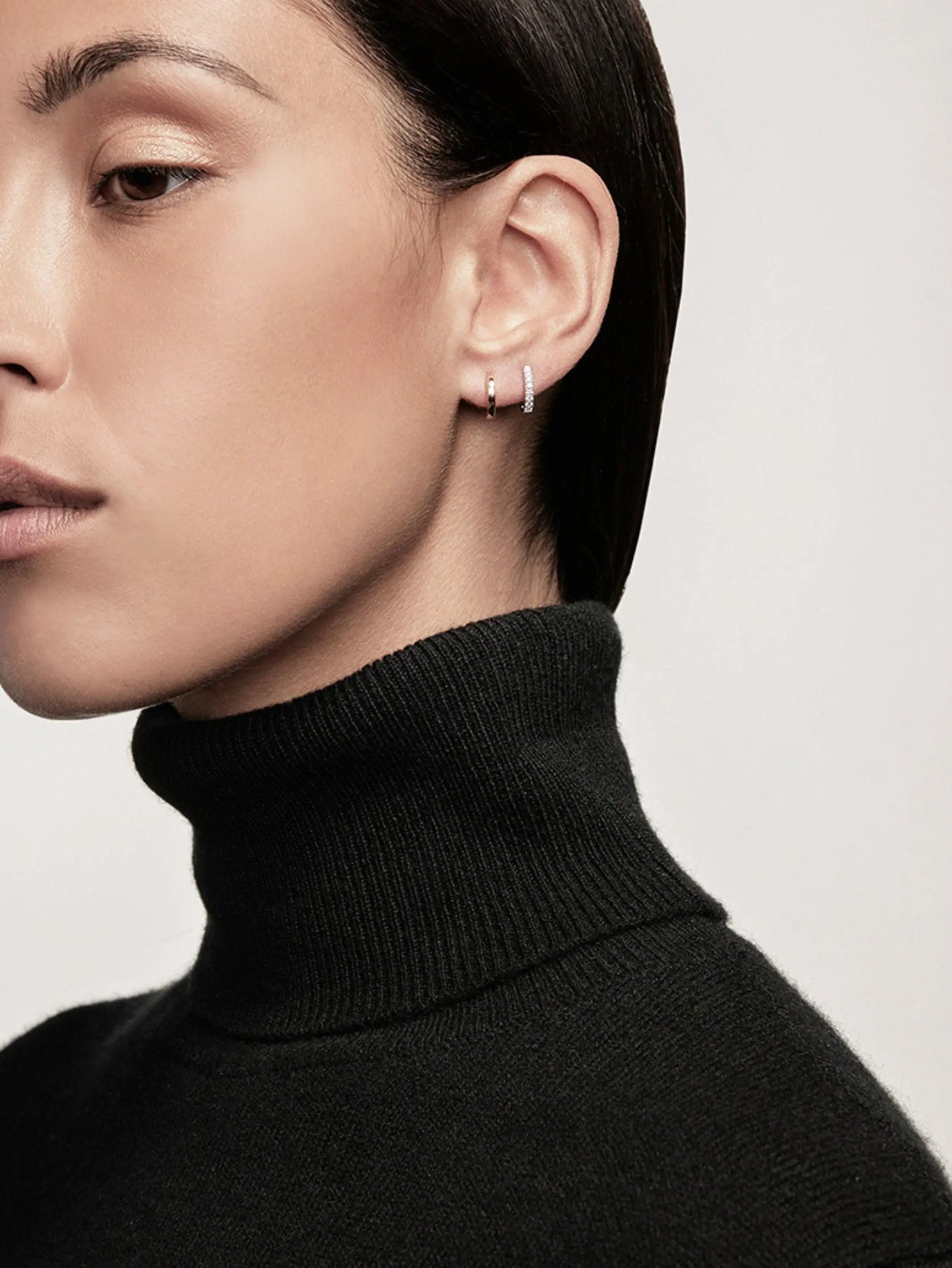 A close-up of a person with a minimalist, stylish appearance. They are wearing a black turtleneck sweater and Huggie Earrings in 18k Rose Gold with post fastening. Their short, dark hair is sleek and neatly combed back. The photograph is taken in neutral, soft light, emphasizing simplicity and elegance.