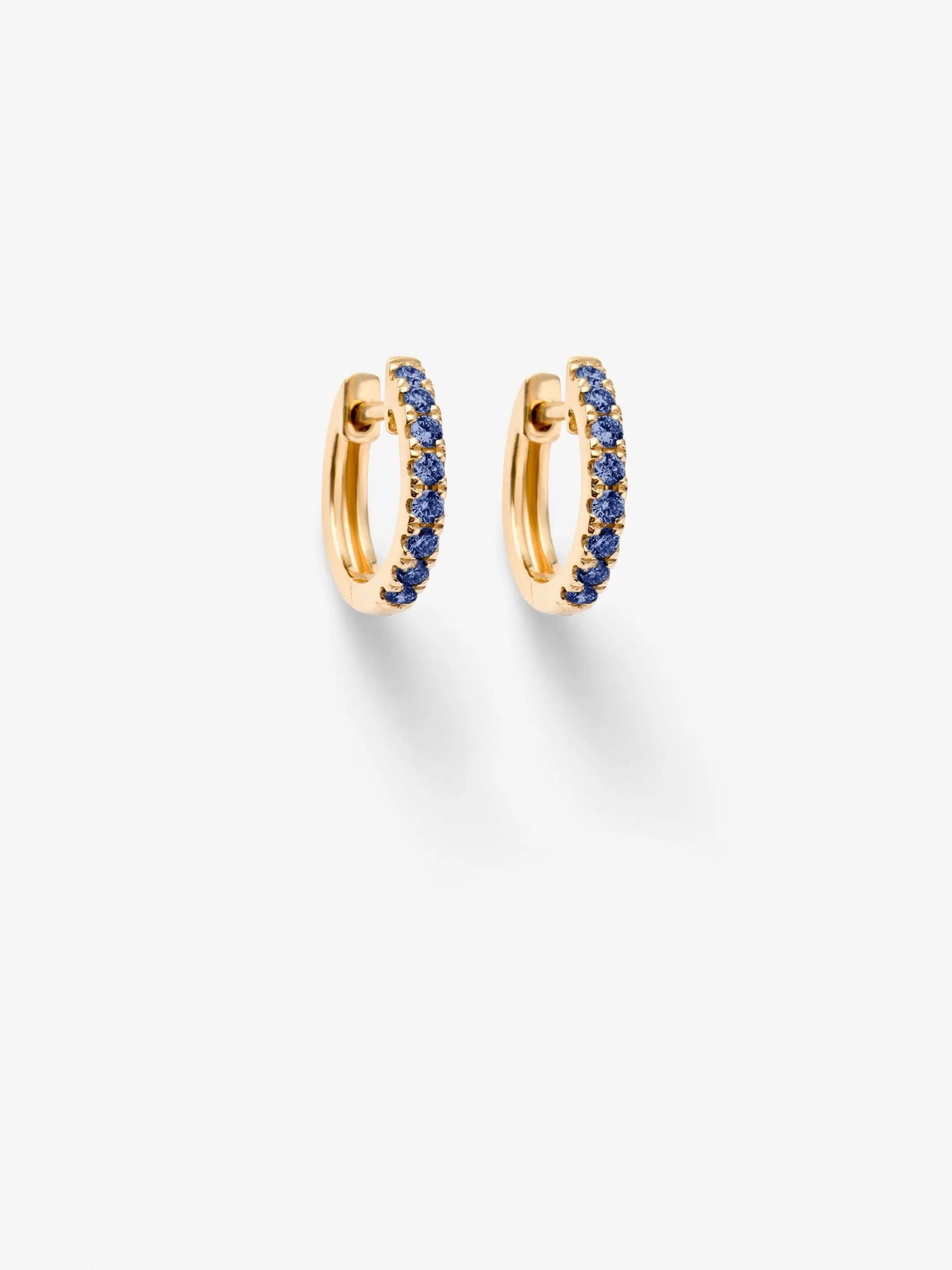 The Huggie Earrings in Blue Sapphire and 18k Gold feature a stylish row of small, blue sapphires embedded along the outer edge of the 18k gold hoops. Presented against a white background, these earrings exude a clean and elegant look. Shadows cast below each earring add depth to their appearance.