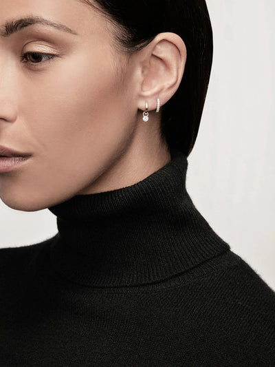 A close-up of a woman wearing a black turtleneck sweater. She has straight, dark hair pulled back and is adorned with two small Huggie Earrings in Diamonds and 18k White Gold in her left ear, one of which features a dangling round gem. The background is plain and light-colored.