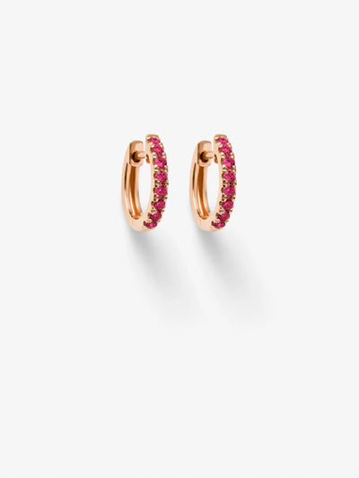 A pair of Huggie Earrings in Ruby and 18k Rose Gold is displayed against a white background. The elegant smaller hoops, encrusted with vibrant pink gemstones, are positioned symmetrically, casting faint shadows beneath them.