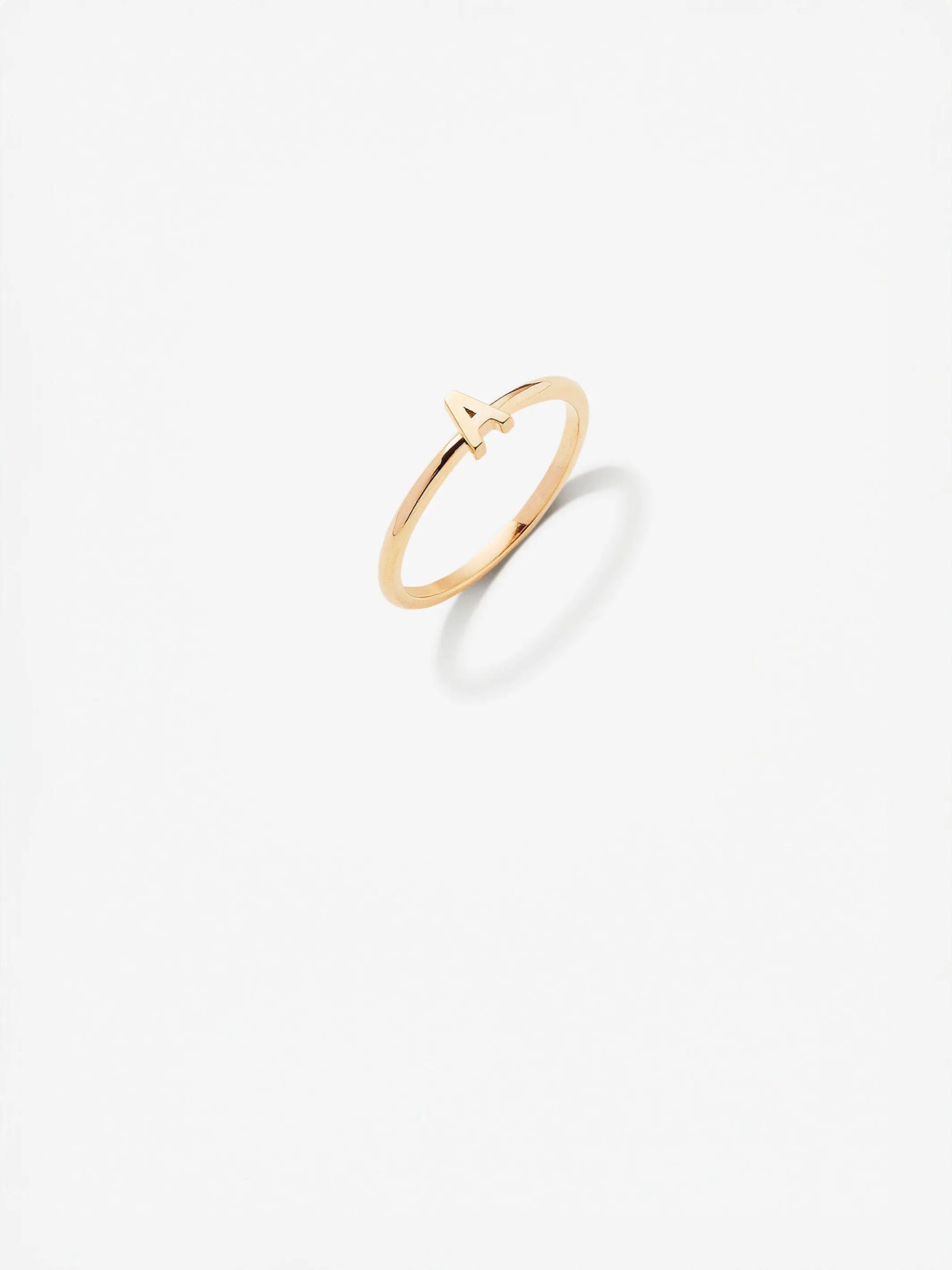 The Letter A Ring in 18k Gold is a delicate, custom-made piece with a minimalistic design that features a small, stylized "A" on top. The 18k solid gold ring casts a soft shadow on a plain white background.