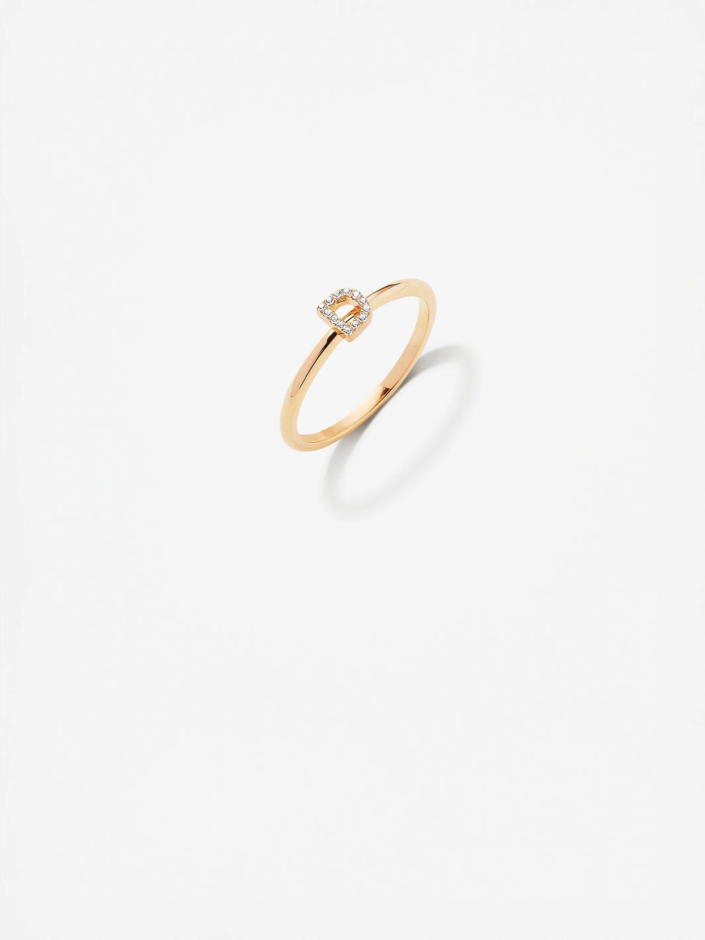 Presenting the exquisite Letter D Ring, crafted in 18k gold with a minimalist band design. This elegant piece showcases a small, square-shaped cluster of radiant diamonds at its center. The ring is displayed on a plain white background to highlight its sophisticated beauty.