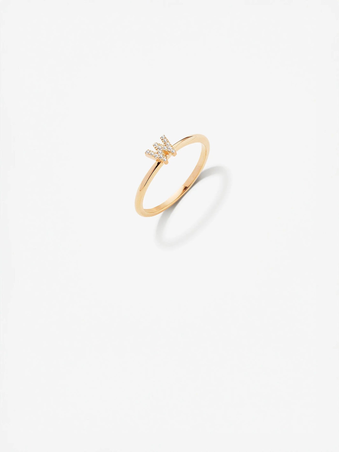 The Letter W Ring in Diamonds and 18k Gold showcases a custom-made design with a delicate 18k gold band. The central "W" shape is adorned with small, sparkling diamonds, all presented against a plain white background that casts a subtle shadow.