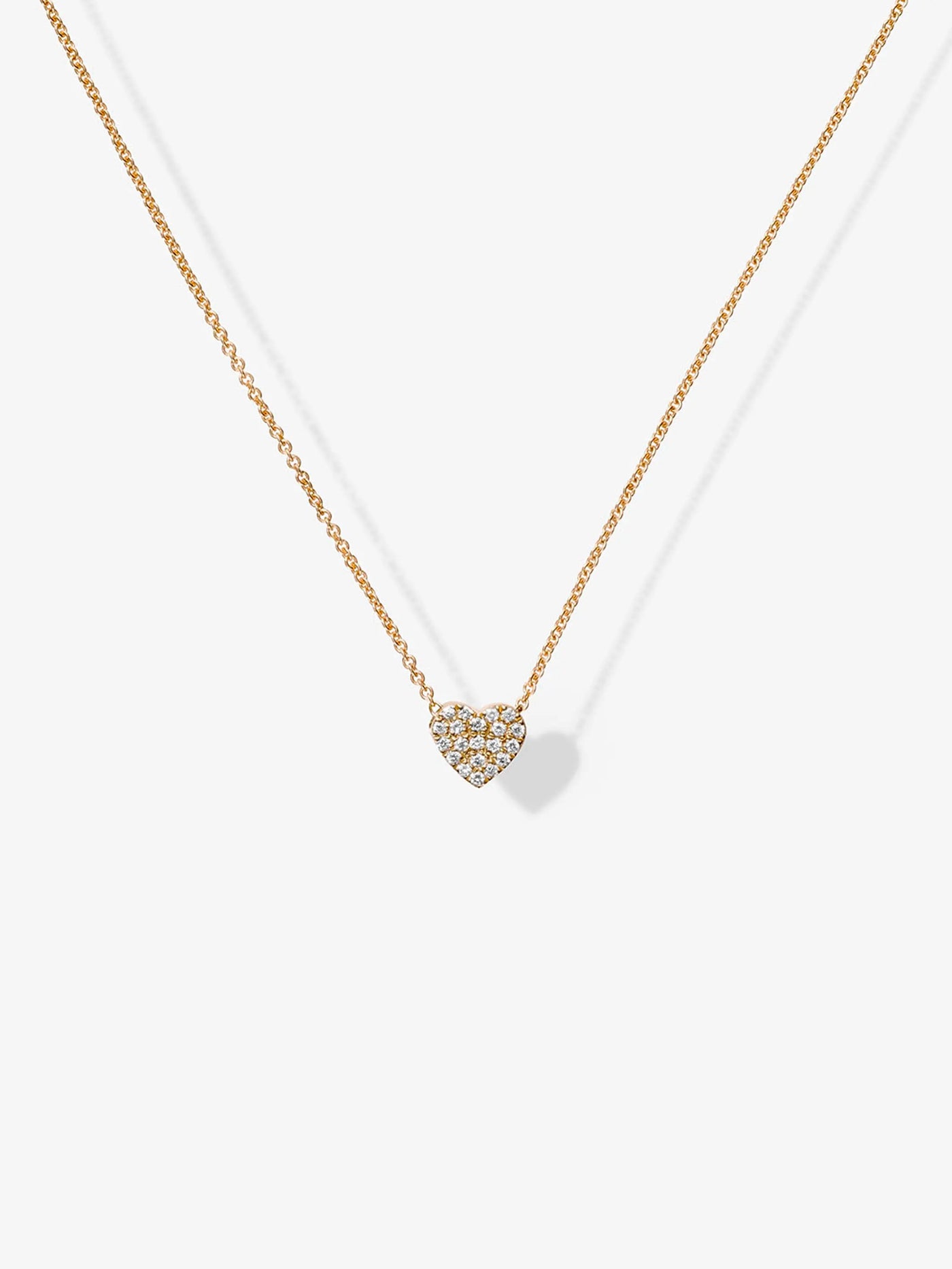 The One Diamond Heart Necklace in 18k Gold showcases a minimalistic design, with a delicate chain featuring a small, heart-shaped pendant encrusted with tiny sparkling crystals. The simple elegance accentuates the shimmering diamond heart against a plain white background.