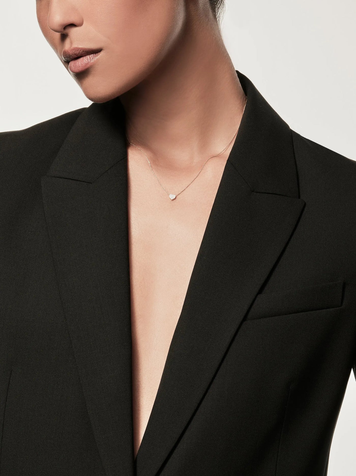 A person wearing the One Diamond Heart Necklace in 18k Gold paired with a deep V-neck black blazer. The image captures the individual from the shoulders up, highlighting the elegant simplicity of both the necklace and blazer against a neutral background.