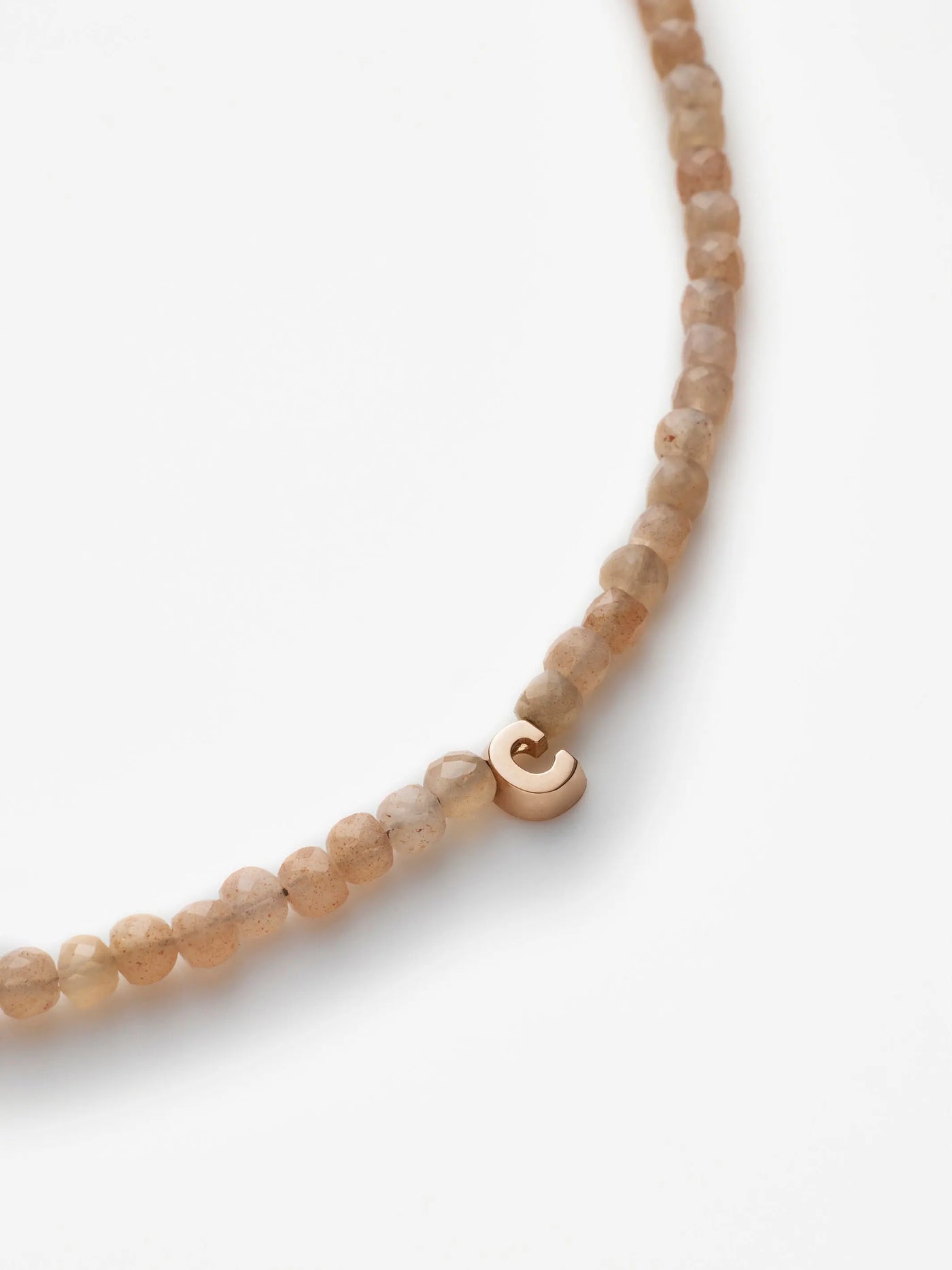 A close-up image of the One Letter Bracelet in Blush Moonstone and 18k Gold, showcasing small, round, light brown blush moonstone beads. The bracelet features a minimalist 18-karat gold clasp shaped like a hook that adds an elegant touch to the piece. The beads are evenly spaced, creating a smooth, seamless appearance.