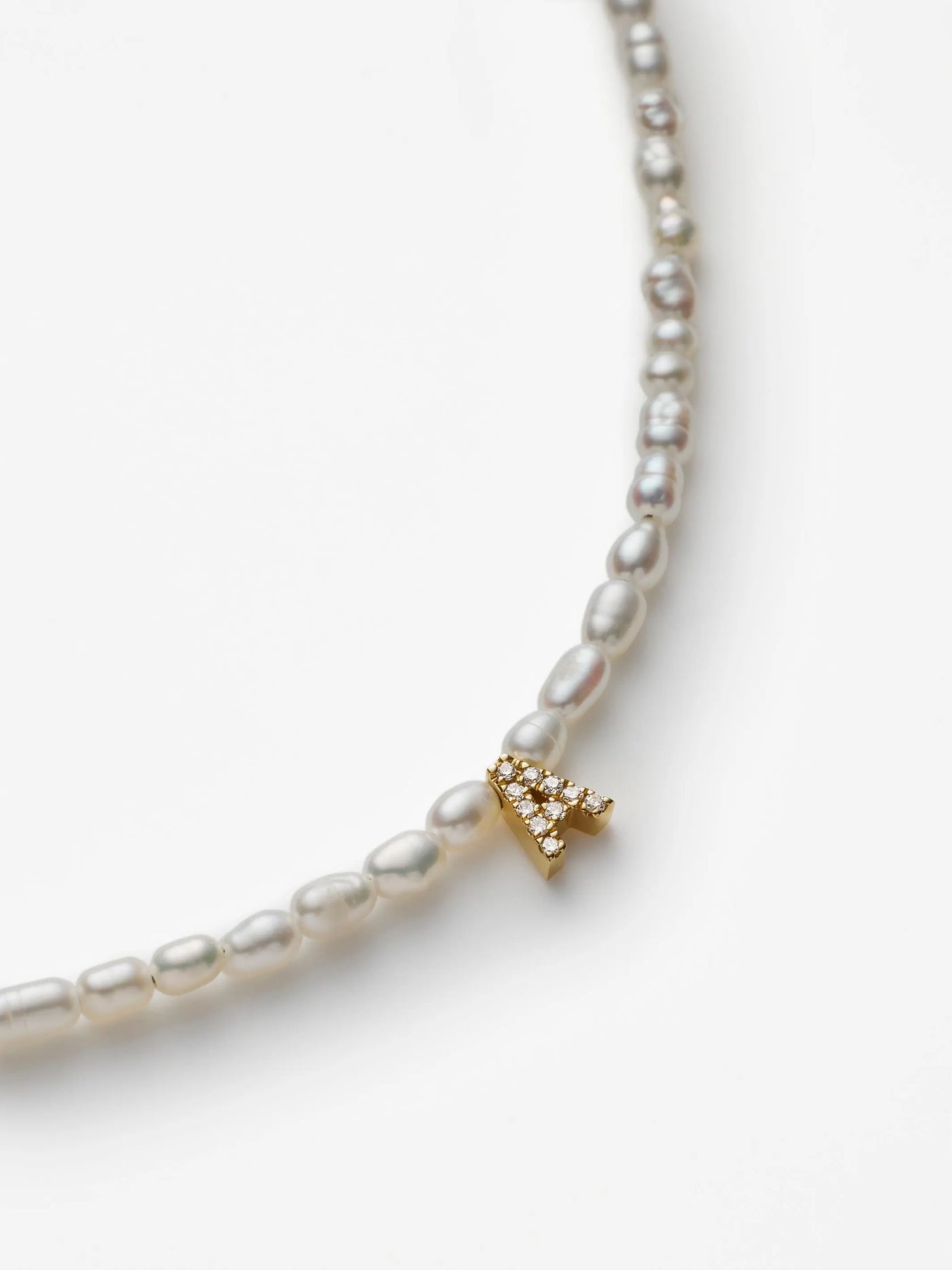 A detailed close-up of the One Letter Bracelet in Diamonds, Pearl, and 18k Gold showcases its elegant design featuring an 18k solid gold charm shaped like the letter 'A,' adorned with sparkling white stones. The pearls display slight variations in size and shape, contributing to a natural and organic aesthetic against a plain white background.