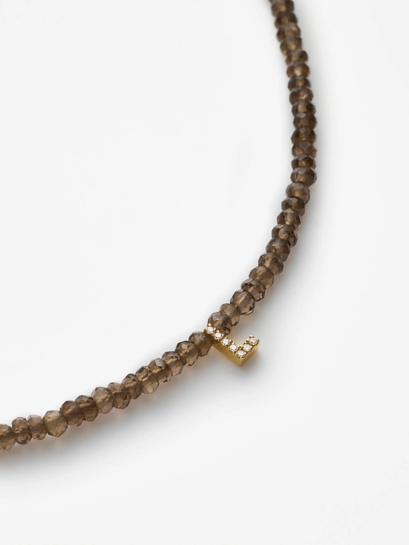 A close-up image of the "One Letter Bracelet in Diamonds, Smoky Quartz and 18k Gold" showcases its hand-strung translucent brown smoky quartz beads. The bracelet is adorned with a small 18k solid gold charm resembling the letter "L," embedded with tiny diamonds that add a touch of sparkle. Custom sizes are available. The background is plain white.