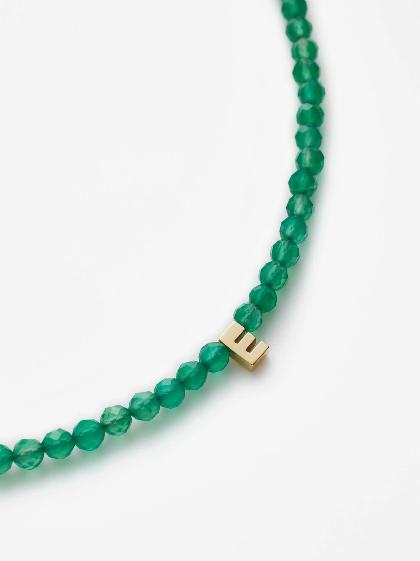 Introducing the One Letter Bracelet in Green Onyx and 18k Gold: a stunning bracelet adorned with green onyx beads and an elegant 18k gold letter "E" charm. This custom-sized accessory is presented against a simple white backdrop.