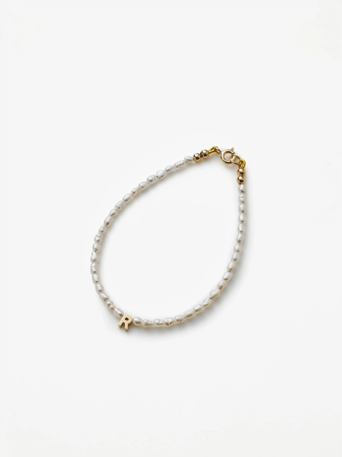 Introducing the One Letter Bracelet in Pearl and 18k Gold, a delicate accessory crafted from small white pearls. It features a single gold letter "R" charm and is available in custom sizes. The bracelet is completed with an 18k gold clasp closure and is presented on a plain white background.