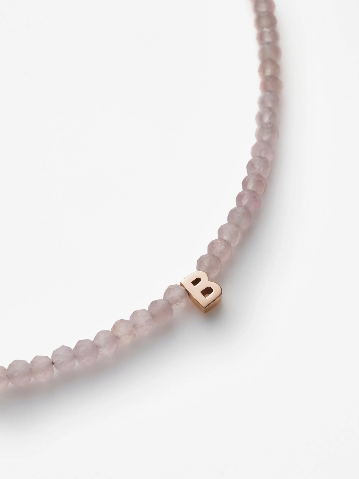 A close-up view of the One Letter Bracelet in Rose Quartz and 18k Rose Gold showcases translucent pinkish beads reminiscent of a rose quartz bracelet, with a single 18k gold letter "B" charm prominently displayed among the beads. The bracelet is set against a white background.