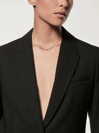 A person is donning a stylish black blazer with a plunging neckline, revealing part of their chest. Around their neck, they wear the One Letter Necklace in Aquamarine and 18k Gold, which adds a touch of elegance with its delicate thin chain. The plain white background draws attention to both the sophisticated clothing and the exquisite jewelry.