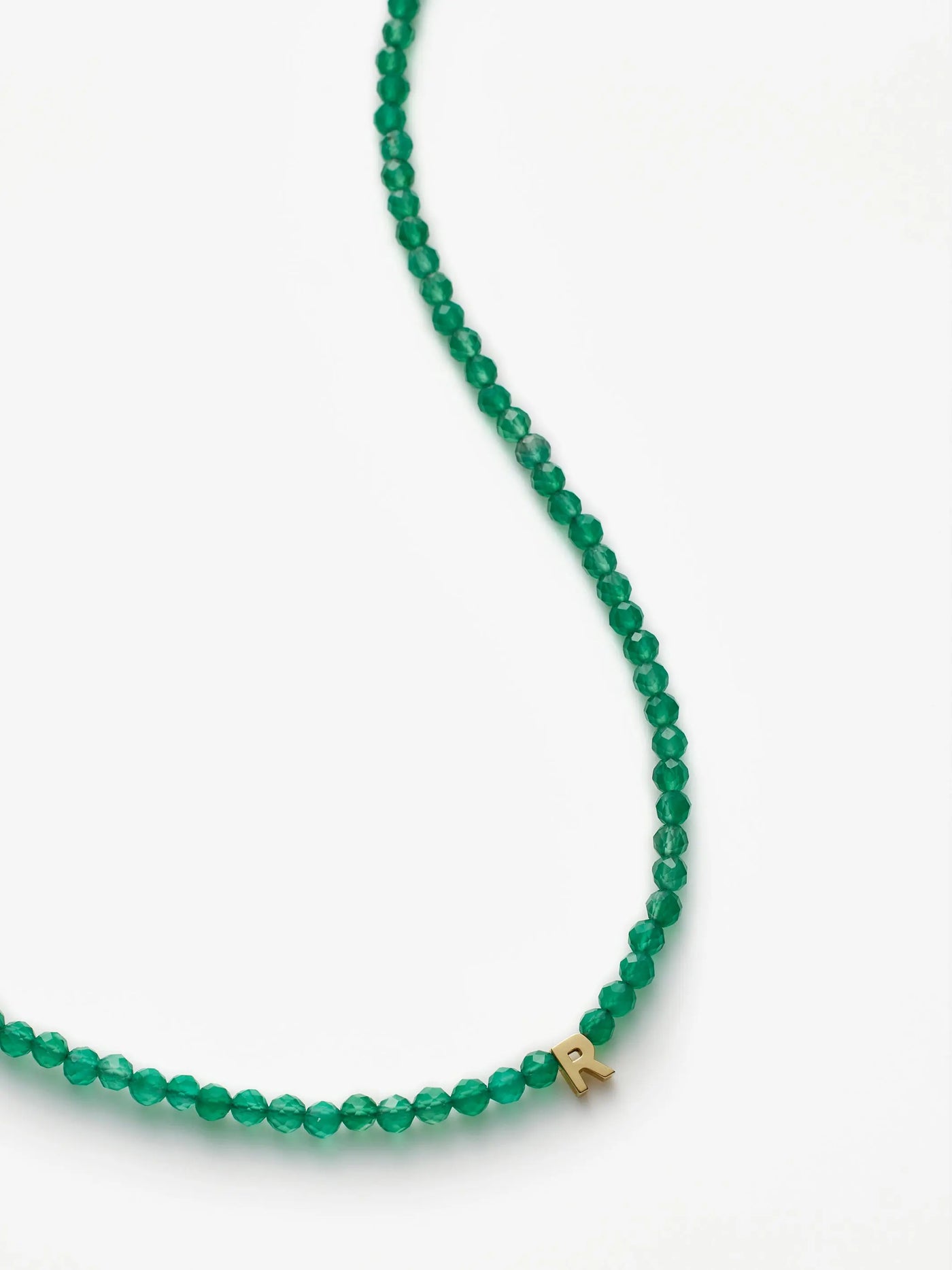 A close-up image of the One Letter Necklace in Green Onyx and 18k Gold, featuring a small gold pendant in the shape of the letter "R" hanging among the green onyx beads. The necklace is set against a plain white background.