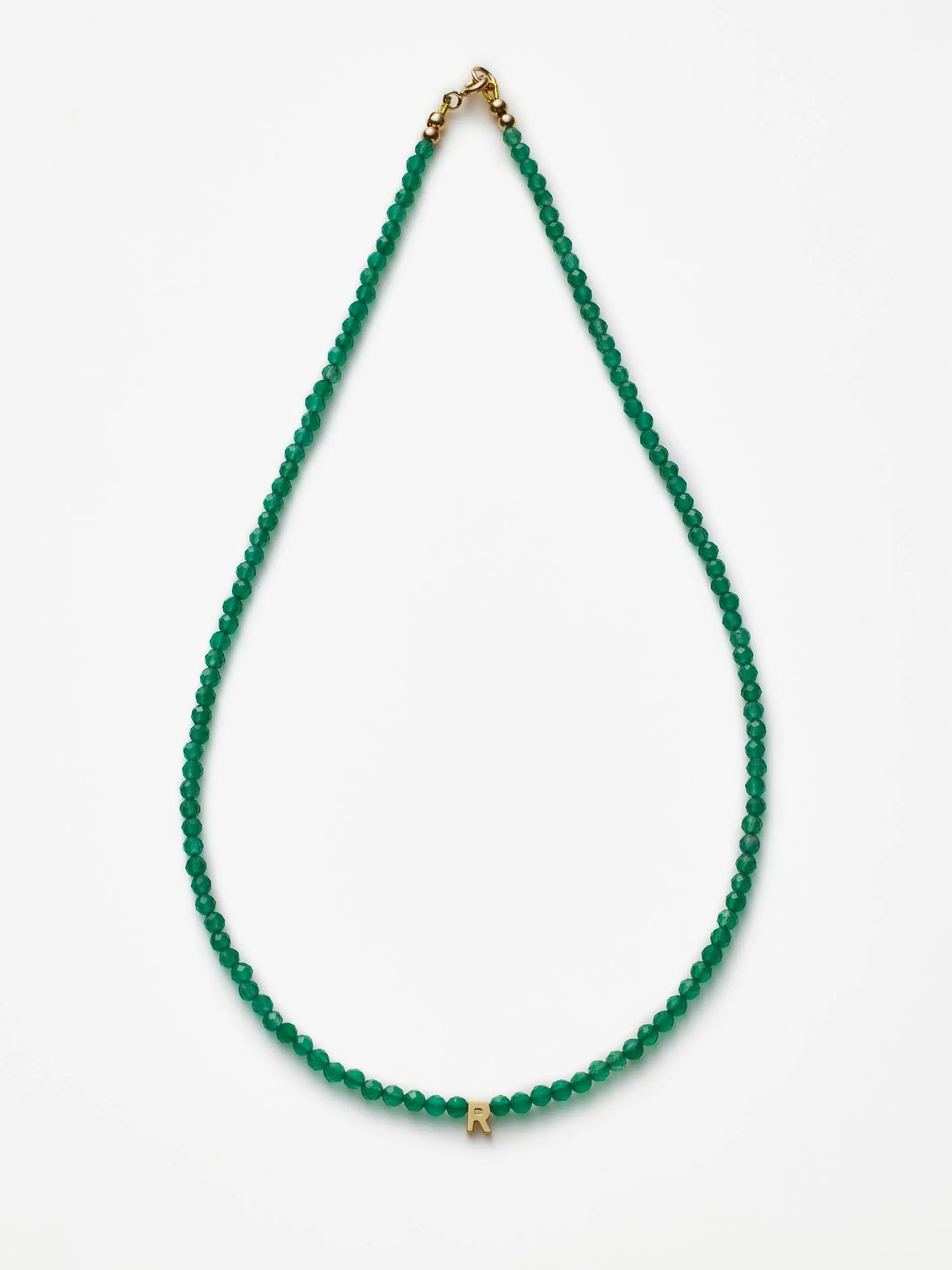 A One Letter Necklace in Green Onyx and 18k Gold, showcasing small green onyx beads with a gold letter "R" pendant in the center, is laid flat against a plain white background. The necklace is completed with an 18k gold-colored clasp at the top.