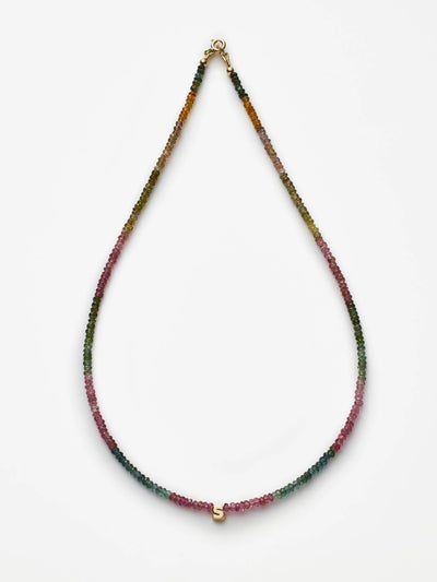 The One Letter Necklace in Tourmaline and 18k Gold is a delicate beaded necklace featuring a gradient pattern of small, multi-colored tourmaline gemstones transitioning from greens to pinks. At its center, the necklace showcases a small 18k gold 'S' charm, set against a plain white background.