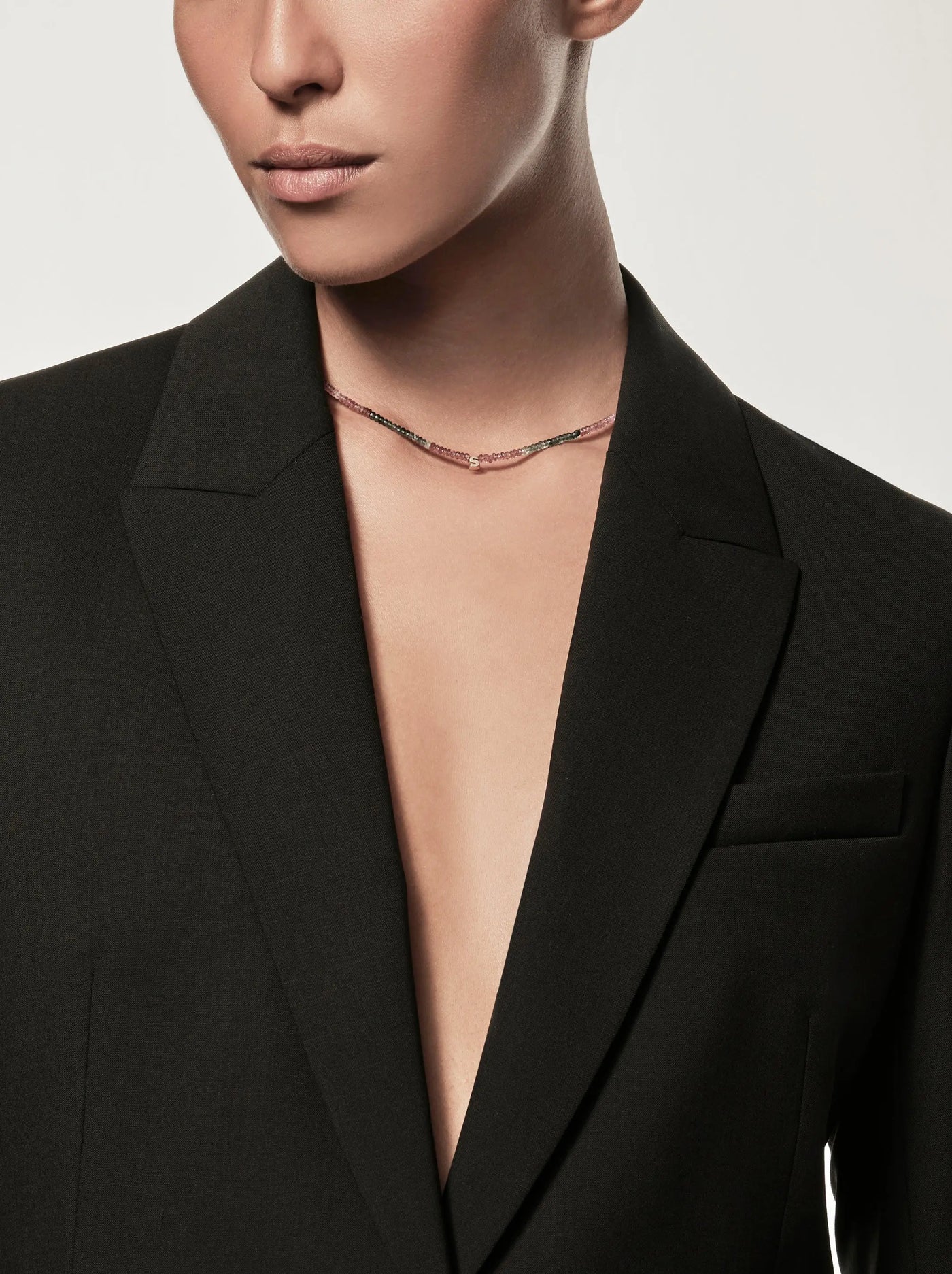 A person with medium skin tone is wearing an open black blazer without a shirt underneath and a delicate One Letter Necklace in Tourmaline and 18k Gold. The background is plain and out of focus, highlighting the attire and accessories. The person's face is partly visible, focusing on their lower half.