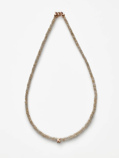 The One Letter Necklace in Zircon and 18k Rose Gold forms a teardrop shape against a plain white background. The necklace features small, translucent beads and a small "K" charm at the bottom center. Natural zircon gemstones add a touch of spiritual balance, while the clasp is visible at the top.