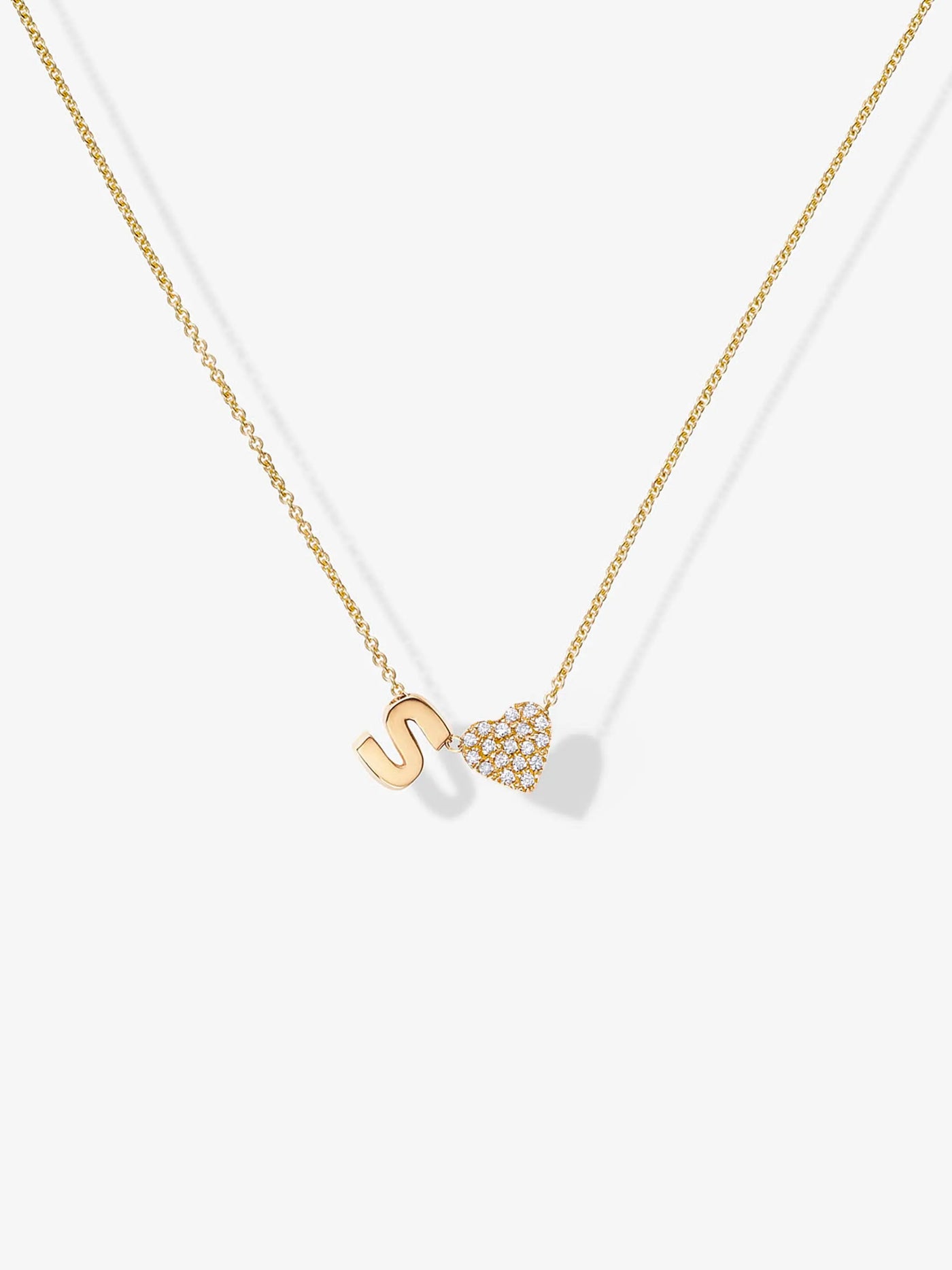 The One Letter and Diamond Heart Necklace in 18k Gold. The image focuses on the torso and neck area, highlighting a minimalist and elegant style. The background is plain and light-colored.