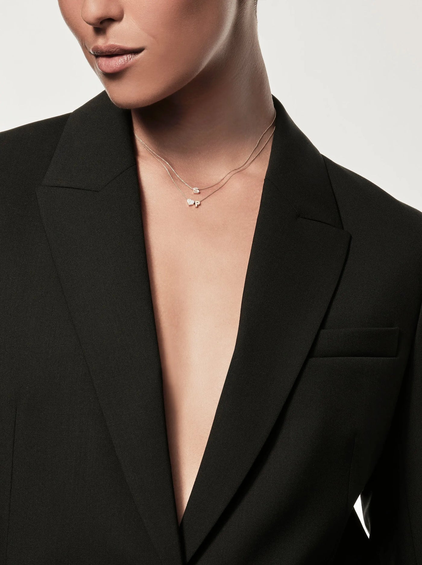 A person wearing a tailored black blazer with an open front, revealing the One Letter and Diamond Heart Necklace in 18k Gold. The image focuses on the torso and neck area, highlighting a minimalist and elegant style. The background is plain and light-colored.