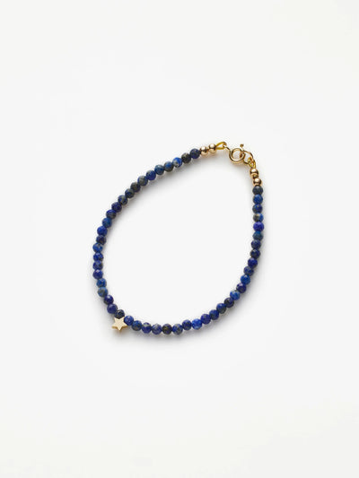 The Star Bracelet in Lapis Lazuli and 18k Gold features delicate, round, faceted blue beads with a single 18k solid gold star charm. This bracelet, embodying wisdom, intuition, and clarity, is secured with a gold clasp closure against a plain white background.