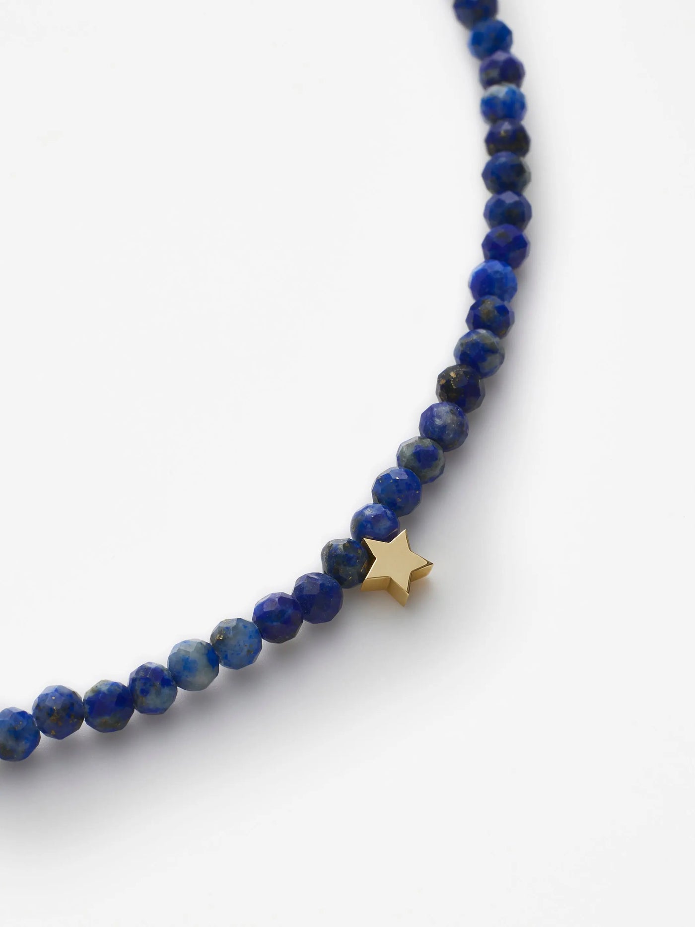 The Star Bracelet in Lapis Lazuli and 18k Gold features delicate, round, faceted blue beads with a single 18k solid gold star charm. This bracelet, embodying wisdom, intuition, and clarity, is secured with a gold clasp closure against a plain white background.